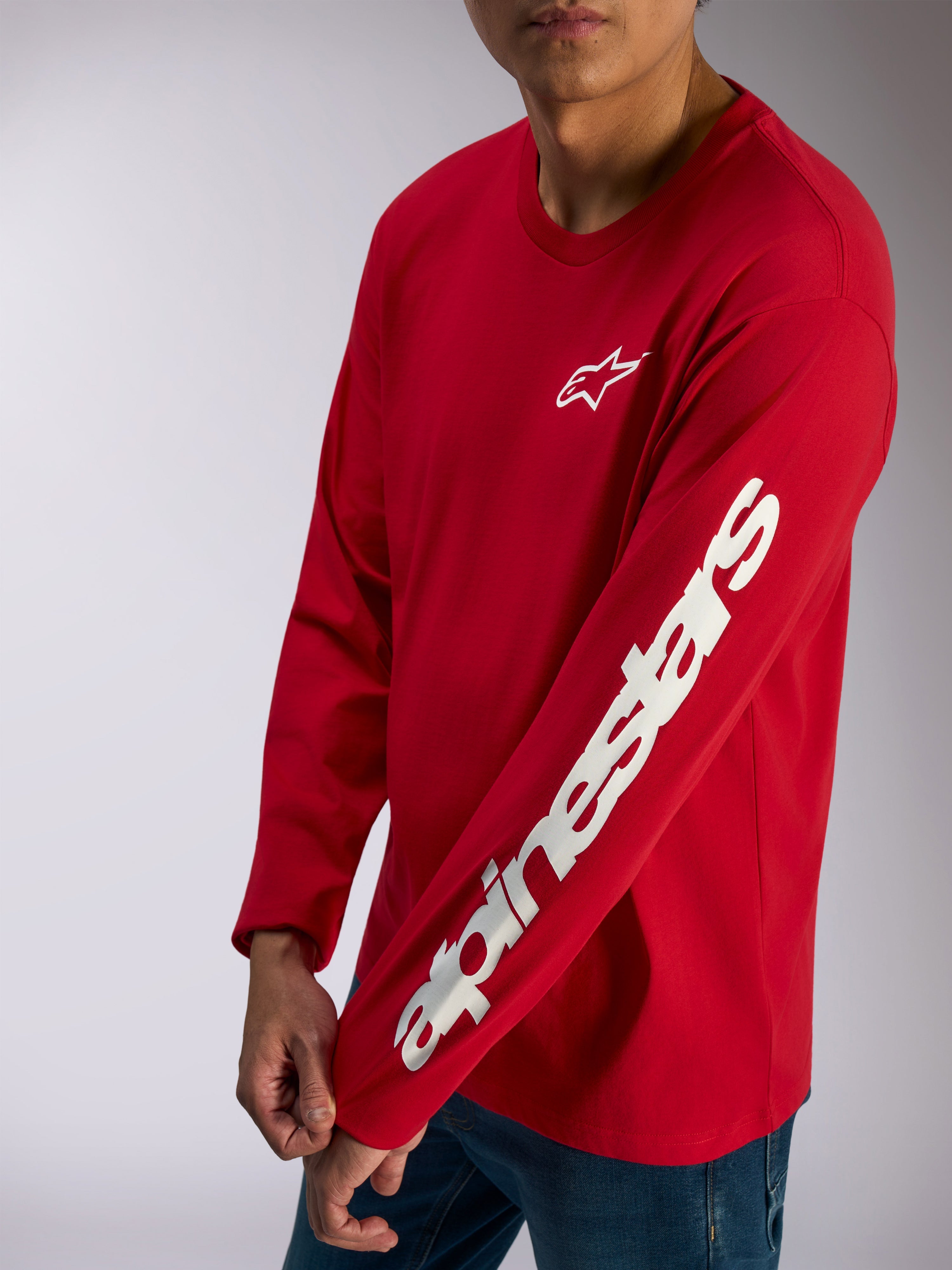 Alpinestars Trackside CSF Tee, Langarm-T-Shirt, Rot, weißes Alpinestars-Logo auf der Brust und markanter Schriftzug auf dem Ärmel, lässige Motorsport-Streetwear kombiniert mit blauen Jeans