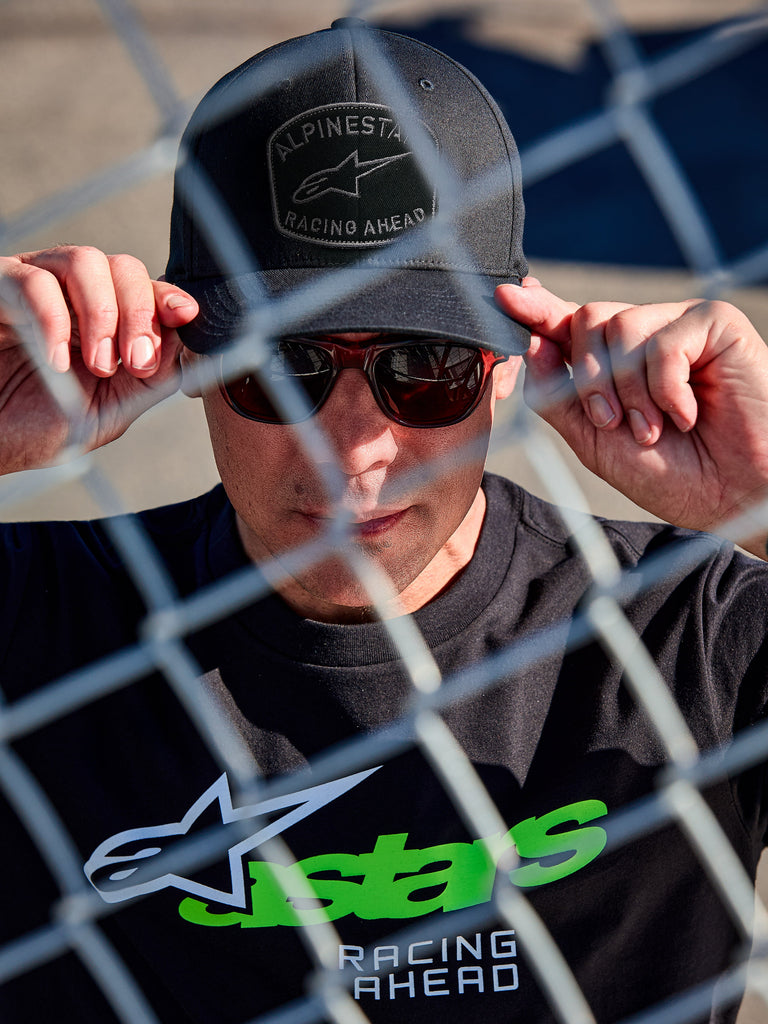 Alpinestars Promptus Snapback Hat, black lifestyle cap, flat bill with structured crown, front curved embroidery patch with braided rope, worn by a man with sunglasses and black t-shirt with green Astars logo, shot through a chain-link fence
