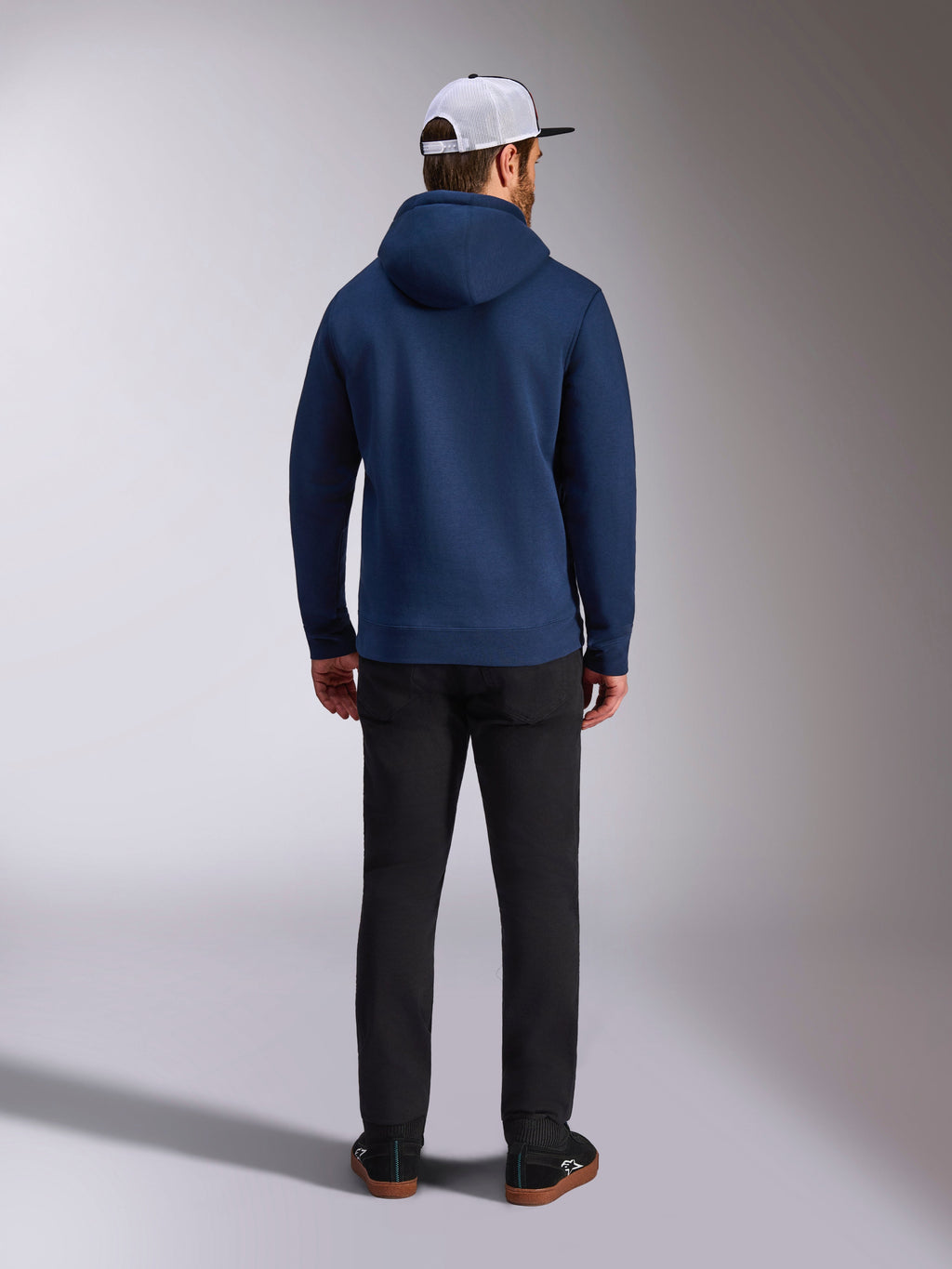 Alpinestars Frontal Hoodie, Marineblau, Rückansicht eines Mannes in einem lässigen Fleece-Kapuzenpullover mit Rippbündchen an Ärmeln und Saum, kombiniert mit einer schwarzen Hose und einer weißen Mesh-Kappe, Motorsport-inspirierte Streetwear