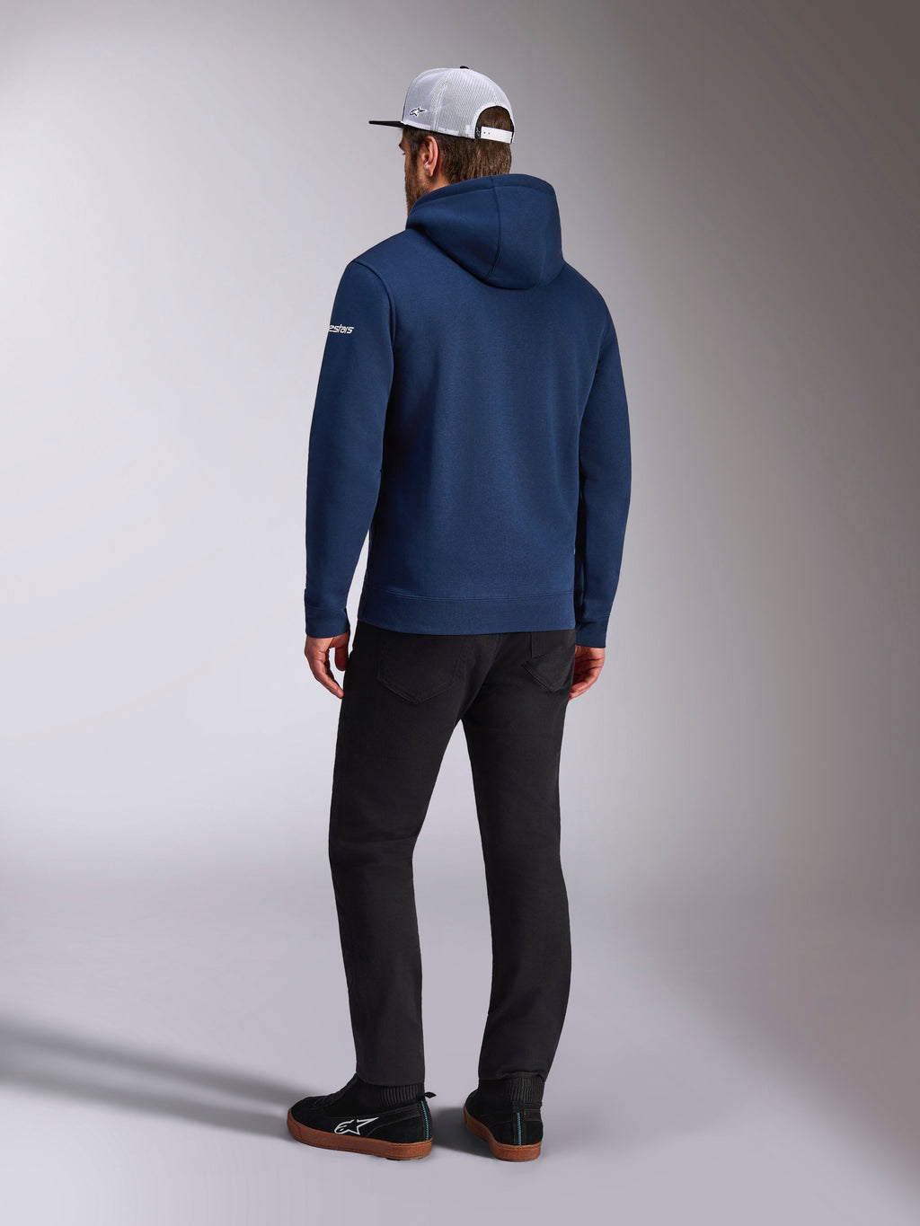 Alpinestars Frontal Hoodie, Rückansicht eines marineblauen Fleece-Kapuzenpullovers, weißes Wortlogo am Ärmel, kombiniert mit schwarzer Hose und weißer Mesh-Trucker-Cap, lässige, vom Motorsport inspirierte Streetwear