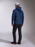 Alpinestars Frontal Hoodie, Rückansicht eines marineblauen Fleece-Kapuzenpullovers, weißes Wortlogo am Ärmel, kombiniert mit schwarzer Hose und weißer Mesh-Trucker-Cap, lässige, vom Motorsport inspirierte Streetwear