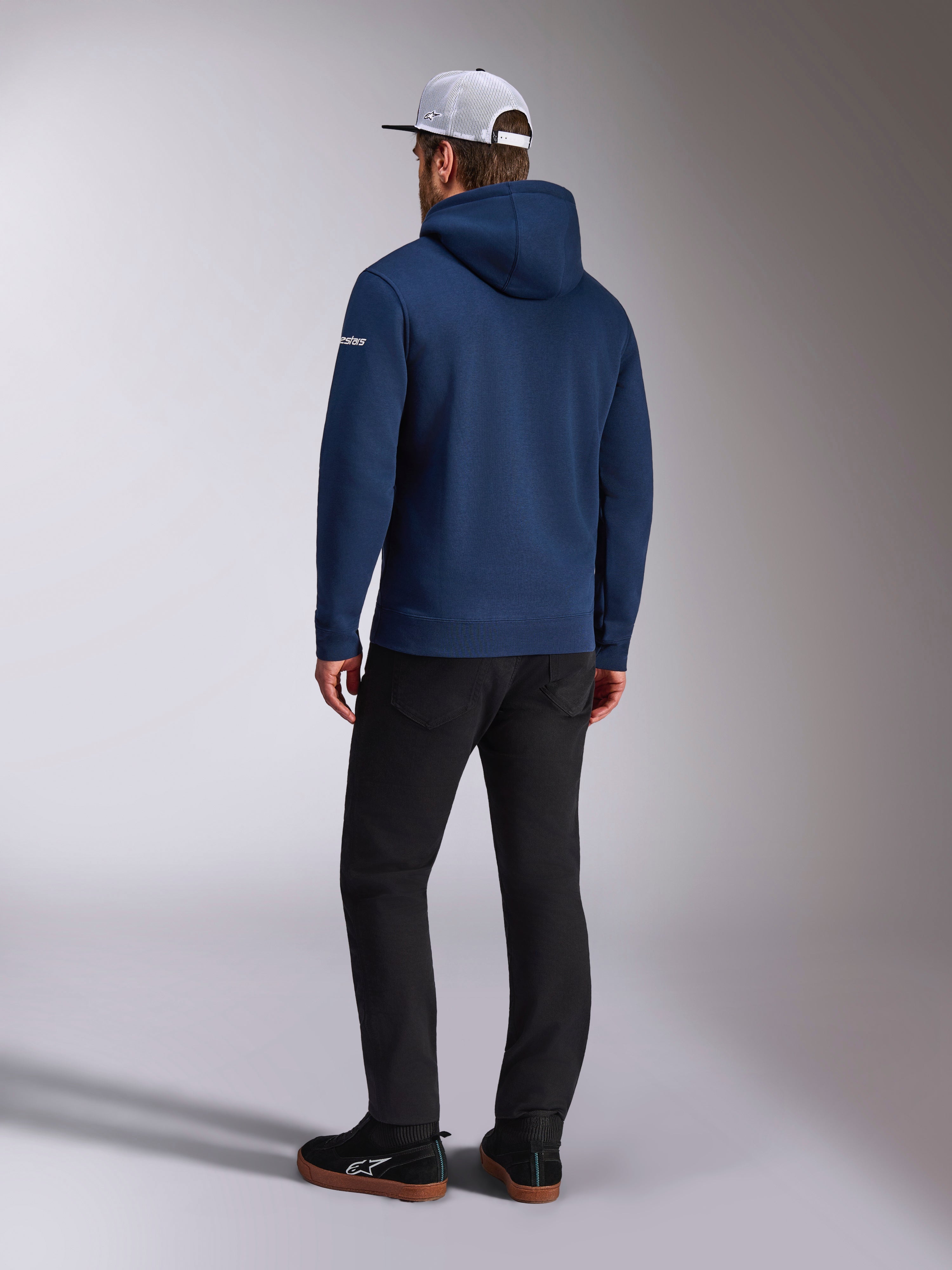 Alpinestars Frontal Hoodie, Rückansicht eines marineblauen Fleece-Kapuzenpullovers, weißes Wortlogo am Ärmel, kombiniert mit schwarzer Hose und weißer Mesh-Trucker-Cap, lässige, vom Motorsport inspirierte Streetwear