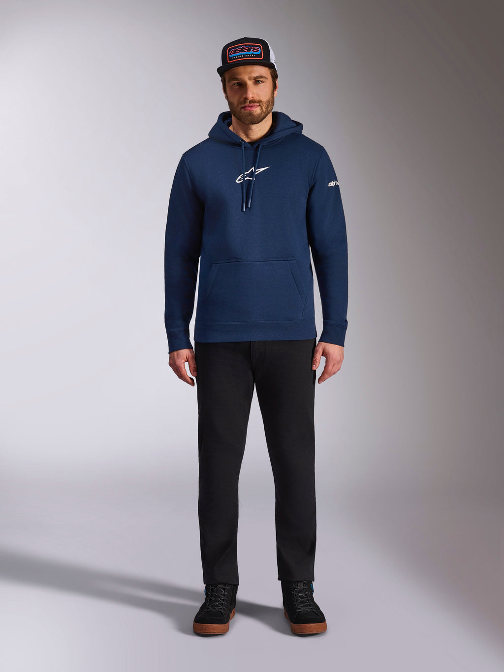 Männliches Model im Alpinestars Frontal Hoodie in Navy, Fleece-Hoodie mit weißem Astars-Logo auf der Brust und Wortlogo auf dem Ärmel, Kängurutasche und Kapuze mit Kordelzug, kombiniert mit einer schwarzen Hose und Markencap, lässige Motorsport-Streetwear
