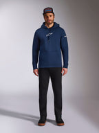 Männliches Model im Alpinestars Frontal Hoodie in Navy, Fleece-Hoodie mit weißem Astars-Logo auf der Brust und Wortlogo auf dem Ärmel, Kängurutasche und Kapuze mit Kordelzug, kombiniert mit einer schwarzen Hose und Markencap, lässige Motorsport-Streetwear