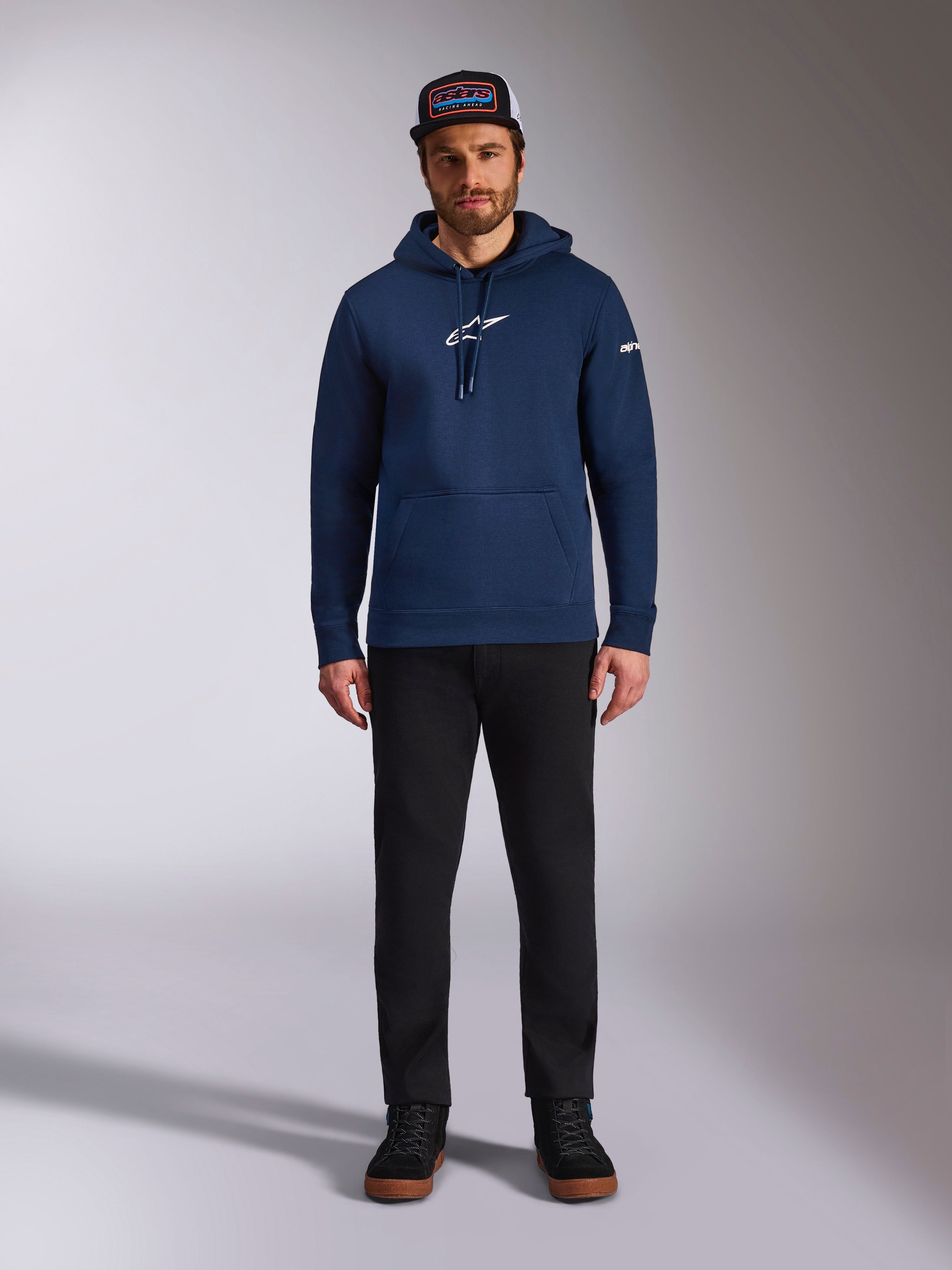 Männliches Model im Alpinestars Frontal Hoodie in Navy, Fleece-Hoodie mit weißem Astars-Logo auf der Brust und Wortlogo auf dem Ärmel, Kängurutasche und Kapuze mit Kordelzug, kombiniert mit einer schwarzen Hose und Markencap, lässige Motorsport-Streetwear