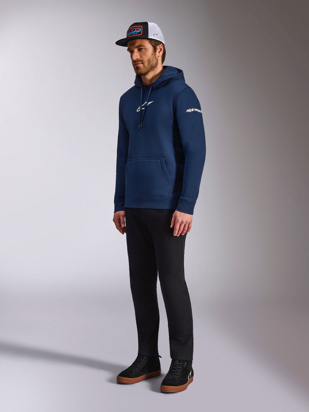 Alpinestars Frontal Kapuzenpullover in Marineblau, lässiger Fleece-Hoodie mit weißem Astars-Logo auf der Brust und Wortlogo auf dem Ärmel, kombiniert mit Trucker-Cap und schwarzer Hose, moderne Motorsport-Lifestyle-Bekleidung