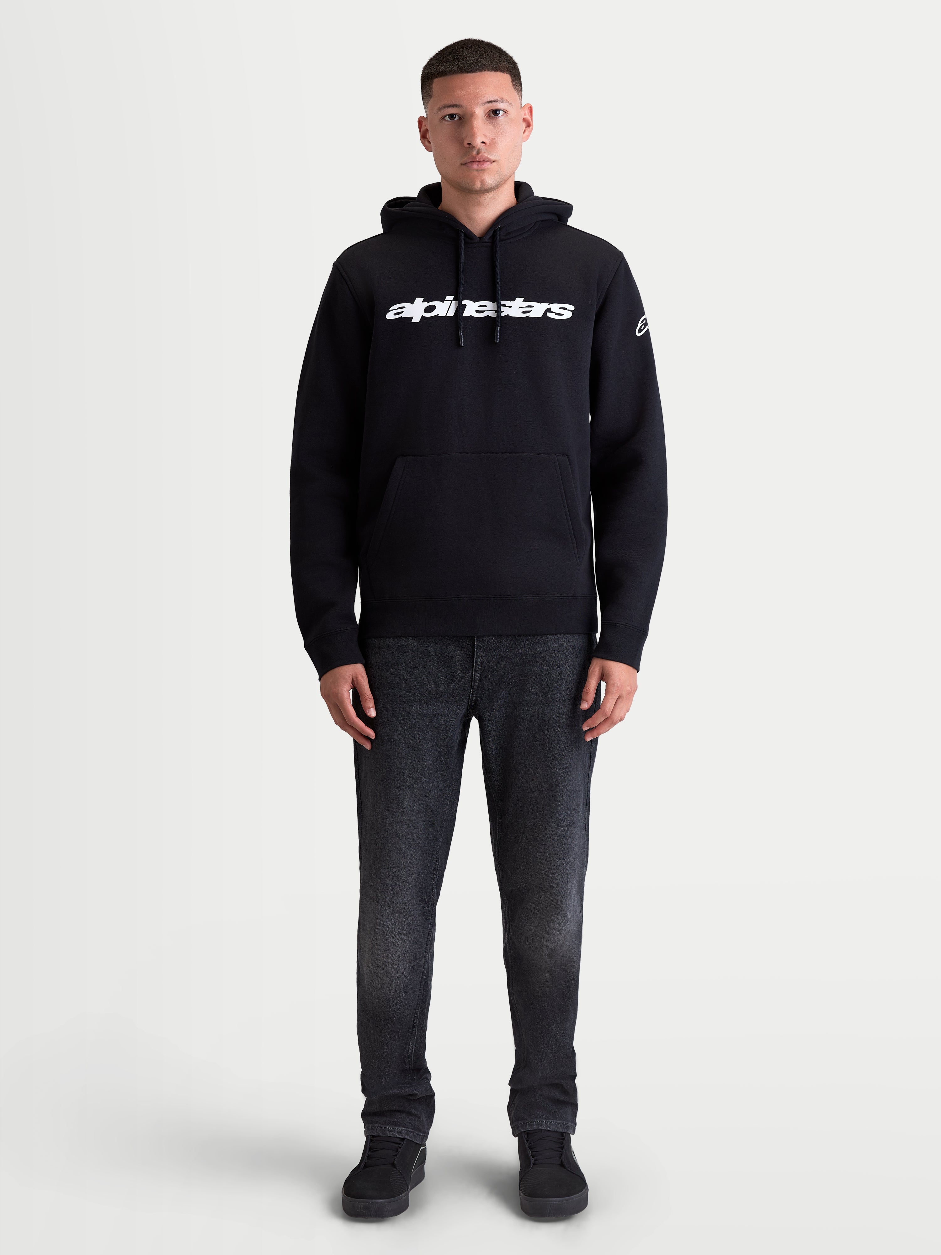 Alpinestars Race Wordmark Hoodie, Sportswear-Kapuzenpullover, Unisex, Schwarz und Weiß, mit Race Linear Word Logo auf der Brust, Ageless-Grafik am Ärmel, Kängurutasche und Kapuze mit Kordelzug, Fleece aus Baumwoll-Mischgewebe, getragen von einem Model.