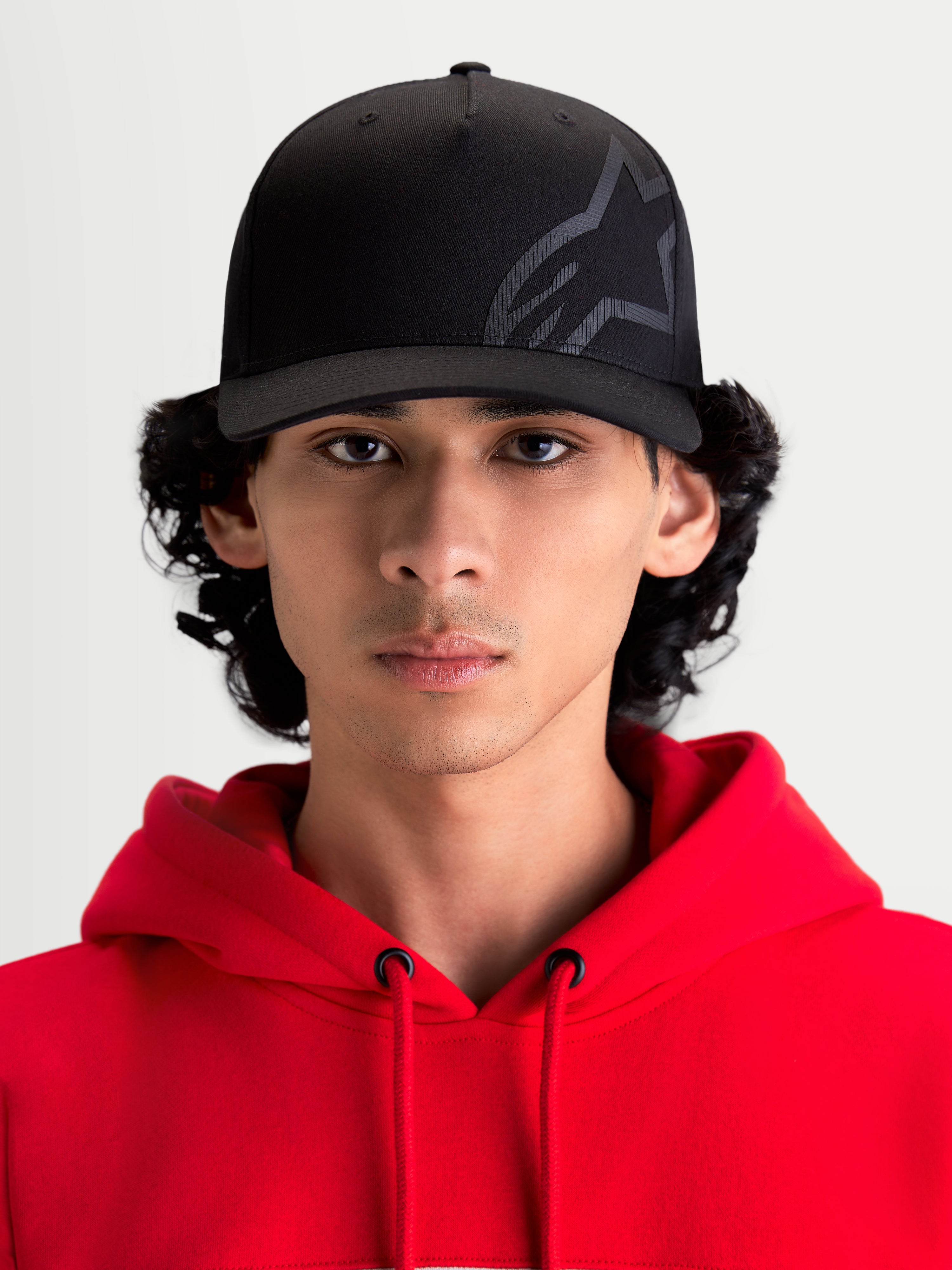 Alpinestars Maze Kappe, schwarze motorsportinspirierte Sportswear-Kappe, Vorderansicht an einem Model mit rotem Kapuzenpullover. Mit strukturierter Krone, gebogenem Schirm und strukturiertem Astars Corp Shift Logo auf strapazierfähigem Twill-Gewebe für einen lässigen Off-Bike-Stil.