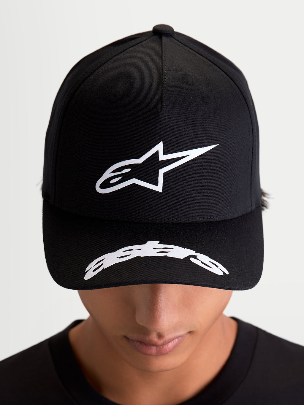 Alpinestars Arclight Kappe, schwarze Sportswear-Baseball-Kappe, Vorderansicht am Modell mit weißem Astars-Logo im Puff-Druck auf der strukturierten Krone und gebogenem Schriftzug auf dem Schirm, motorsport-inspirierte Freizeitkleidung.