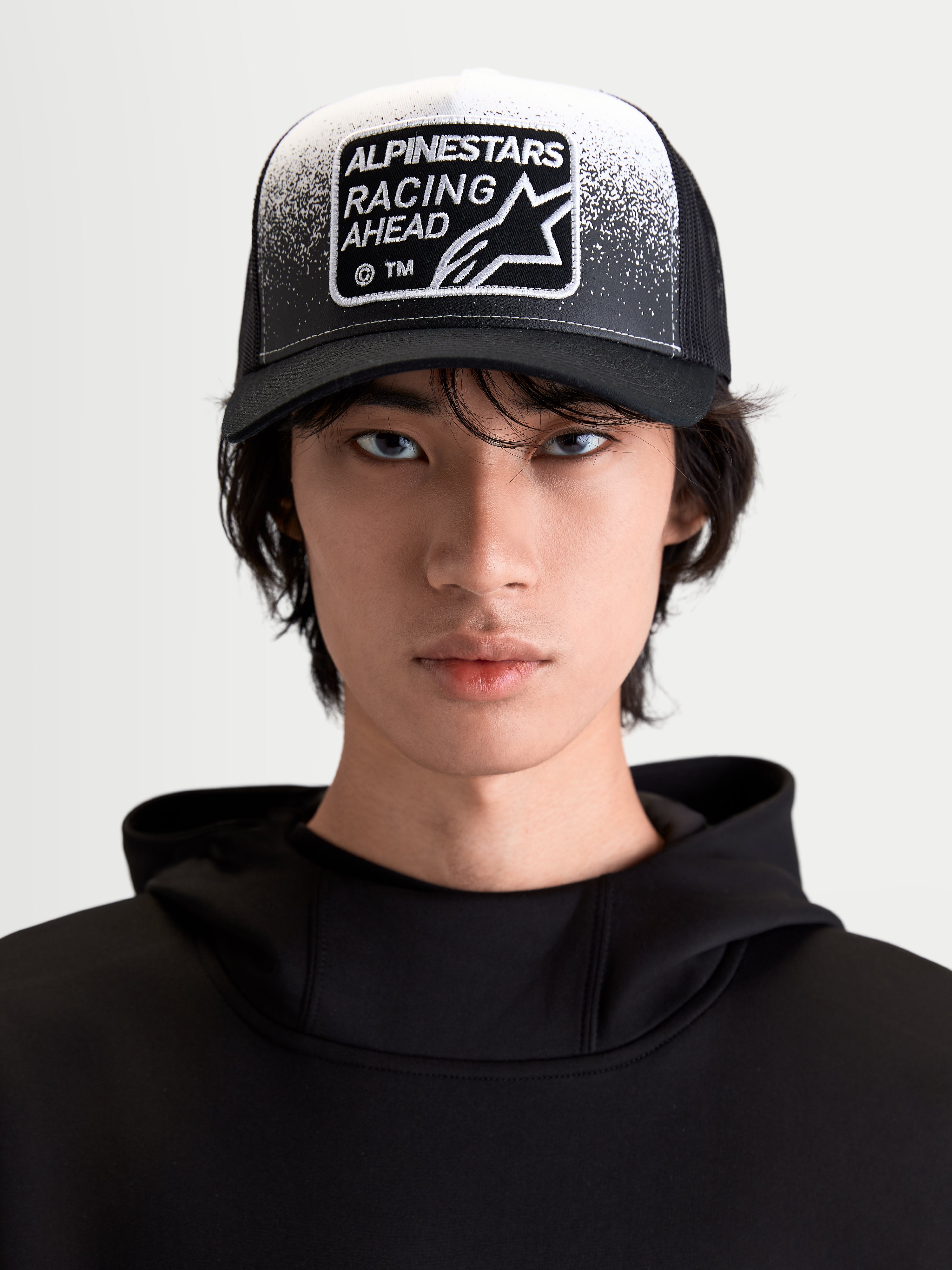 Alpinestars Gravel Trucker-Kappe, Sportswear-Lifestyle-Cap für Herren und Damen, Schwarz und Weiß, Frontansicht am Modell mit gebogenem Schirm, atmungsaktiver Mesh-Rückseite und gewebtem Racing Ahead Patch mit Kieselstruktur-Detail.
