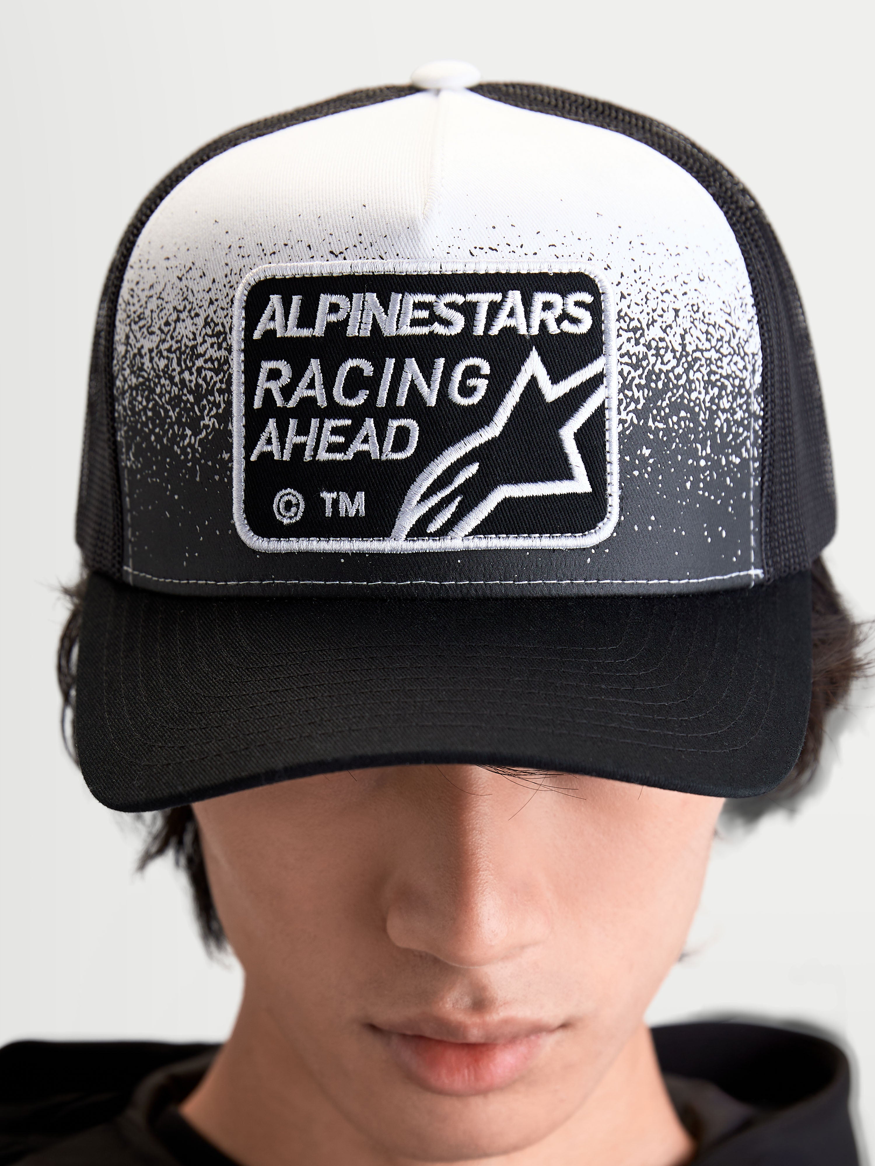 Alpinestars Gravel Trucker Hat, Sportswear Trucker Cap for Men and Women, Black and White, featuring a woven Alpinestars Racing Ahead patch with gravel-texture detailing, structured crown, and curved bill, on-model front view.
