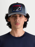 Alpinestars Gravel Trucker-Kappe, lässige Sportswear-Cap mit Mesh-Rückseite für Damen und Herren, Dunkelgrau und Schwarz, an einem Modell gezeigt. Mit strukturierter Krone, gebogenem Schirm und einem gewebten Front-Patch mit Alpinestars Racing Ahead Branding und Kieselstruktur-Details.