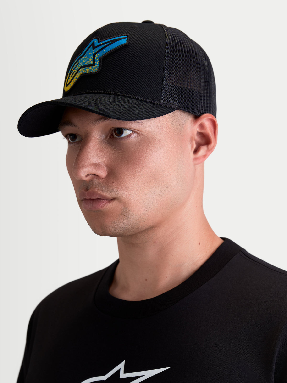 Alpinestars Meteorite Trucker-Kappe, Sportswear Casual Lifestyle-Kappe mit Mesh-Rückseite, Herren, Schwarz, mit einem blau-gelben Astars Logo-Patch in Kieselstruktur, gebogenem Schirm und strukturierter Krone, am Modell getragen.