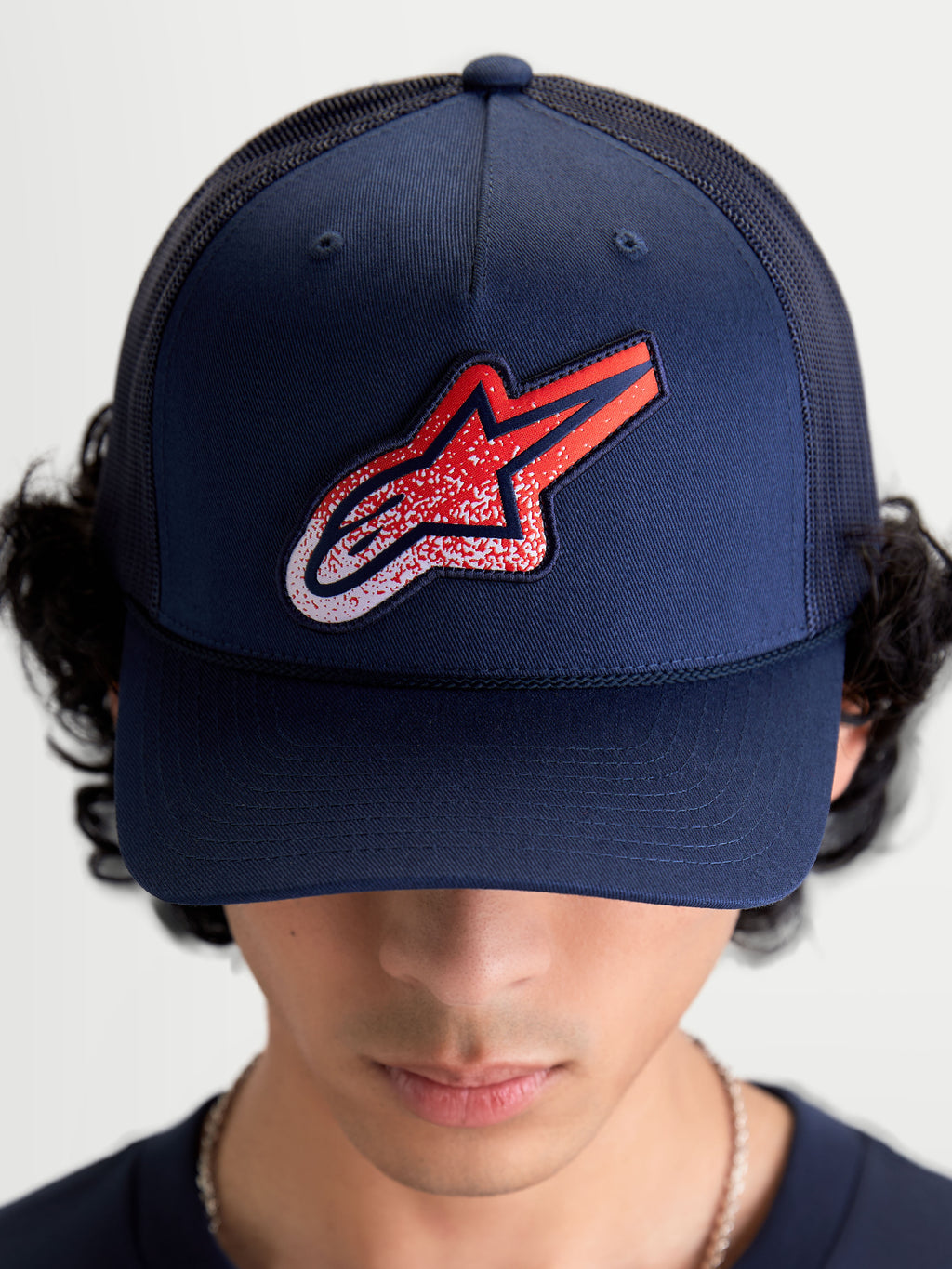 Alpinestars Meteorite Trucker-Kappe, Sportswear Mesh-Kappe, Blau mit rot-weißem Astars-Logo-Patch in Kieselstruktur, strukturierte Krone und gebogener Schirm, Vorderansicht am Modell getragen.