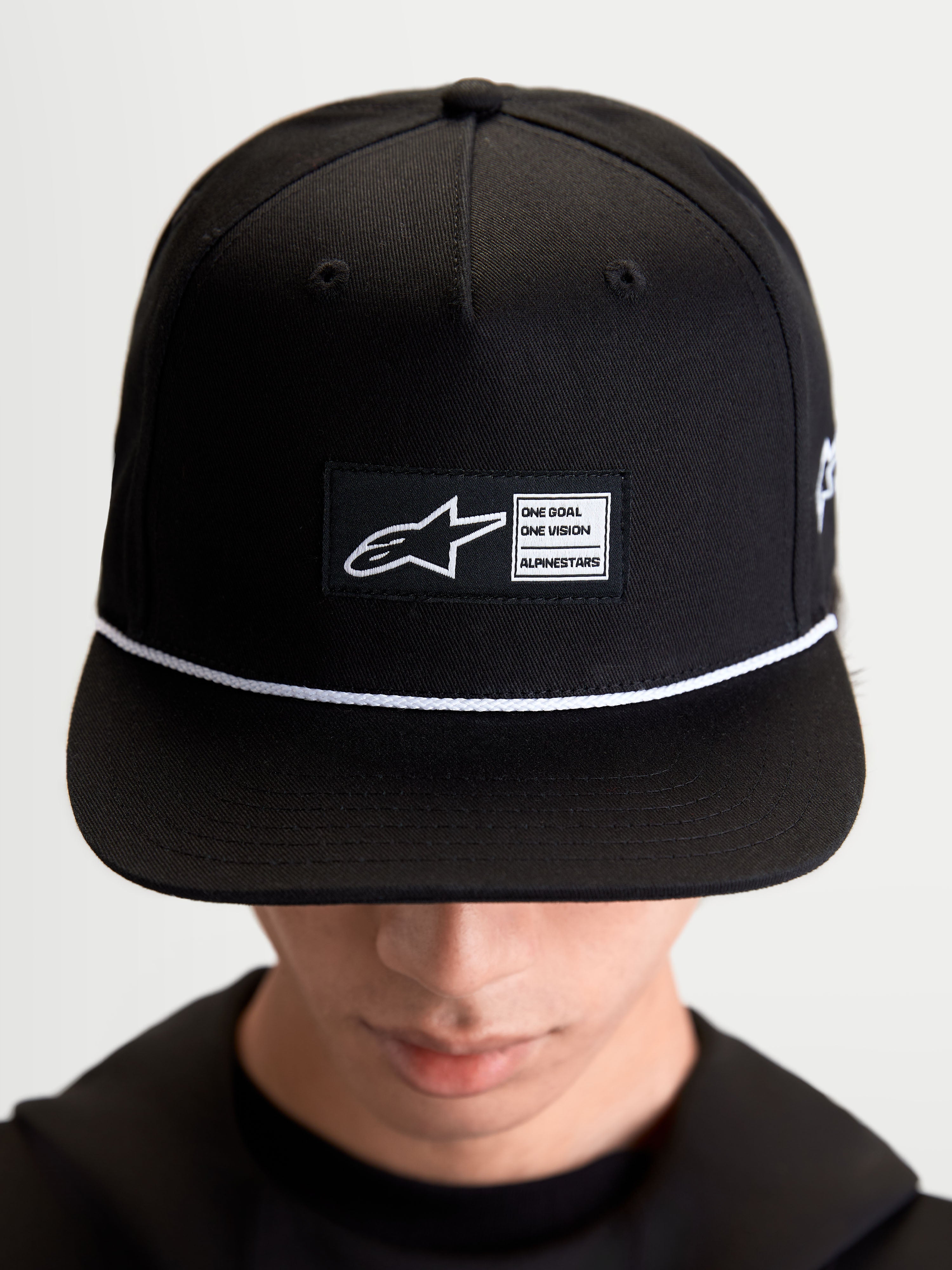 Alpinestars Label Snapback Kappe, Sportswear-Lifestyle-Cap, Unisex, Schwarz und Weiß, aus Baumwolltwill, mit strukturierter Krone, flachem Schirm, weißem Kordelakzent und gewebtem Alpinestars Logo-Patch auf der Vorderseite, am Model getragen.