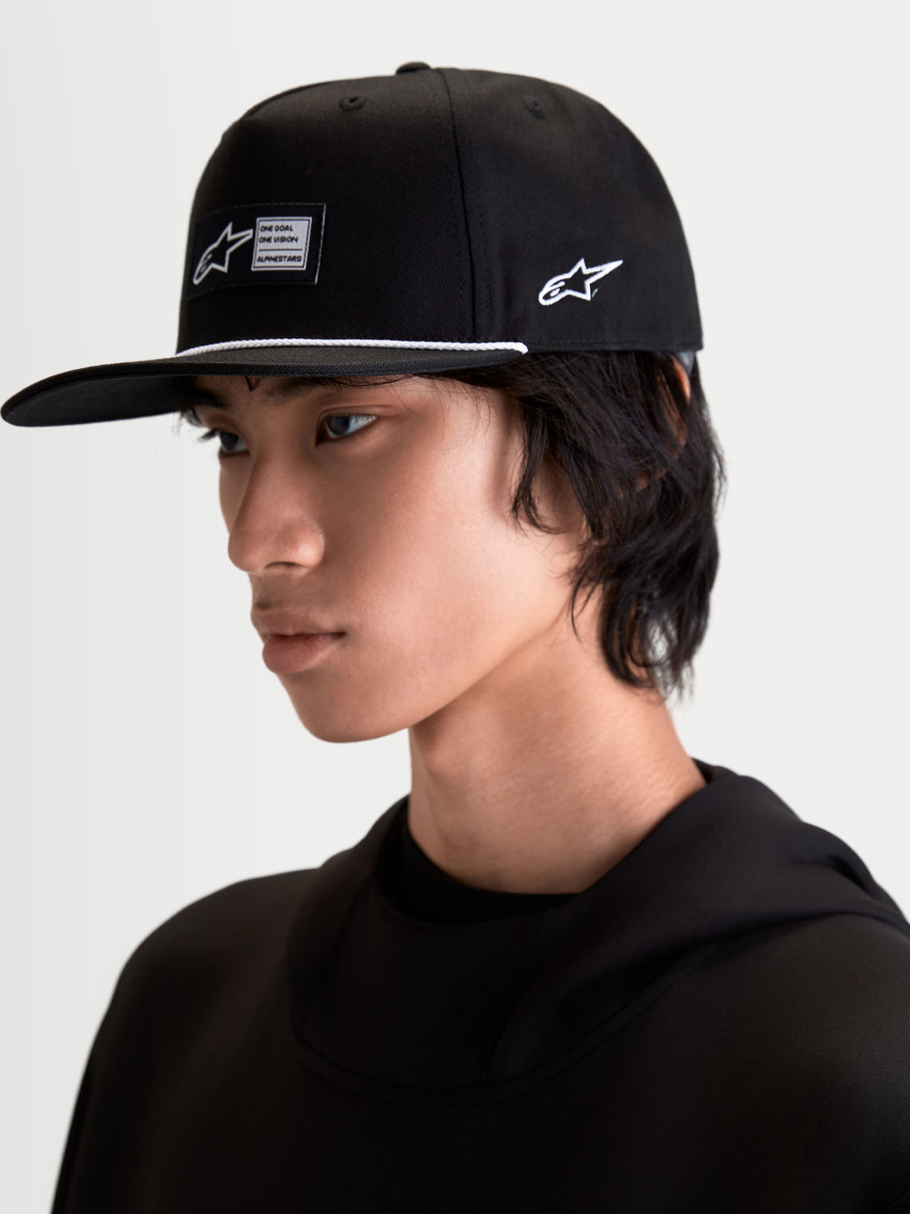 Alpinestars Label Snapback Kappe, schwarze Sportswearkappe mit flachem Schirm und strukturierter Krone, gewebtes Frontlabel, weißes Akzentseil und seitliche Logo-Stickerei, getragen von einem Model für einen modernen Motorsport-Lifestyle-Look.