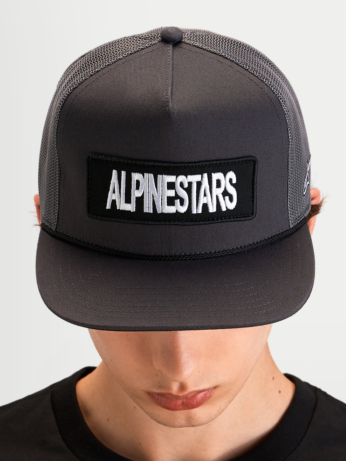 Alpinestars Ten Trucker Hat, sportswear flat-bill cap worn by a model, dark grey, featuring a front Alpinestars word logo patch and structured mesh trucker crown