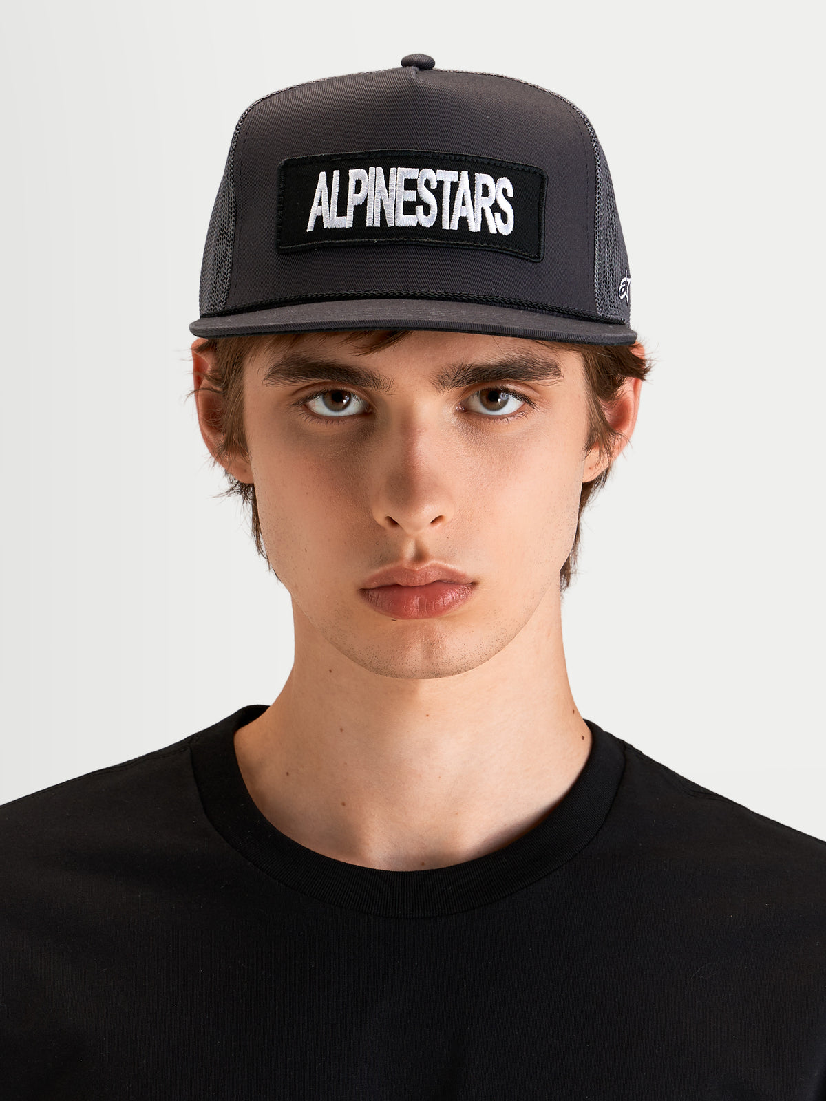 Alpinestars Ten Trucker Hat, flat bill sportswear cap in dark gray, worn by a model, featuring a structured crown and front Alpinestars word logo embroidery patch