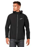 Männliches Model trägt Alpinestars Kascade Regenjacke, Schwarz, weißes Brustlogo und Ärmel-Branding, Regenjacke mit Kapuze, beschichtetem Frontreißverschluss und Klettverschlussbündchen, kombiniert mit dunkler Jeans, lässige Motorsport-inspirierte Streetwear