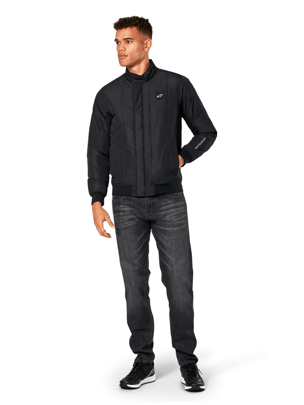 Alpinestars Precedent Jacke, gesteppte Sportjacke, Schwarz, getragen mit dunkelgrauer Jeans und schwarzen Sneakern, mit schrägem Steppmuster, niedrigem Stehkragen, Rippstrickbündchen an Ärmeln und Saum, weißem Logo-Patch auf der Brust und Stickerei am Ärmel.
