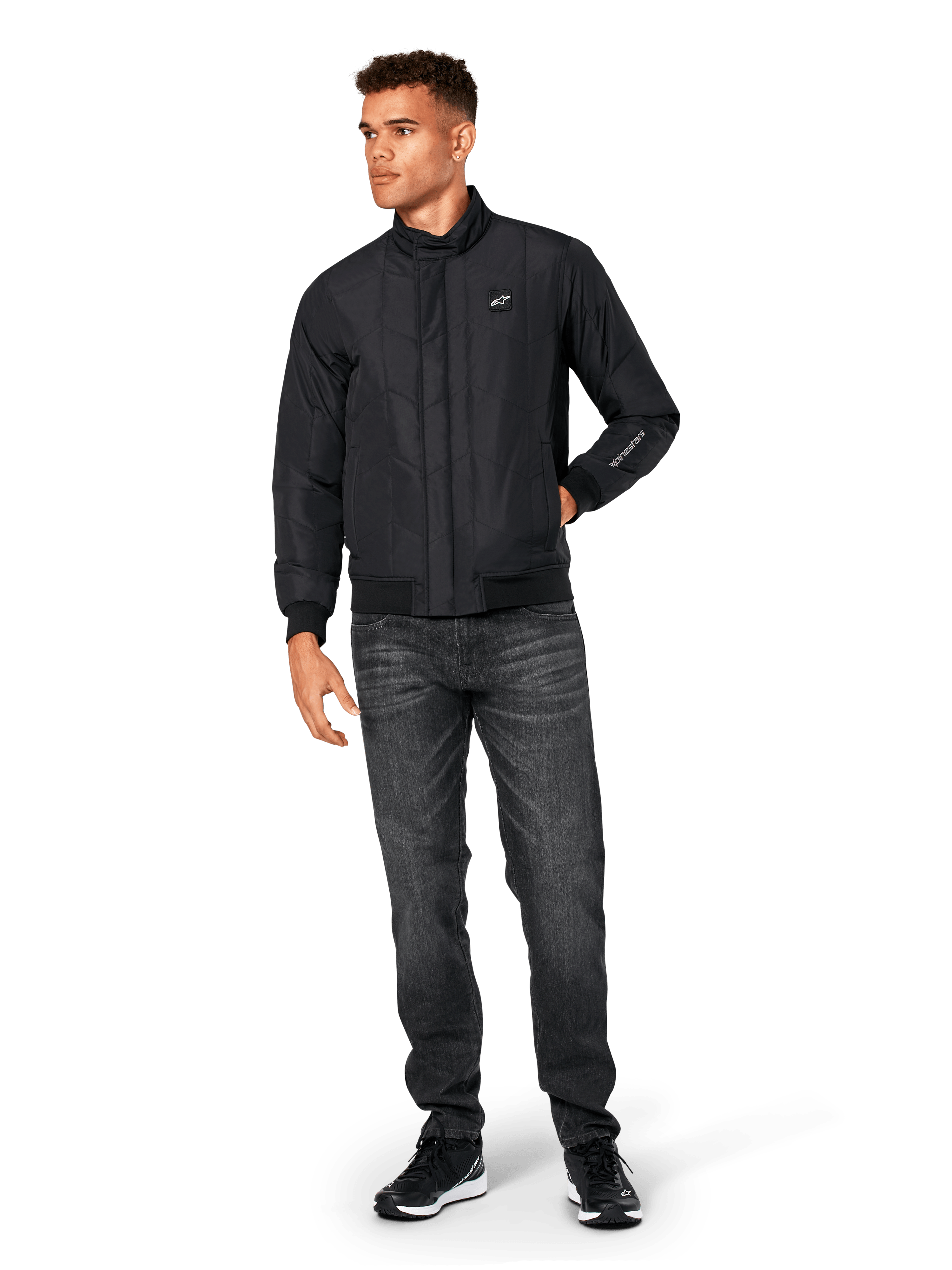 Alpinestars Precedent Jacke, gesteppte Sportjacke, Schwarz, getragen mit dunkelgrauer Jeans und schwarzen Sneakern, mit schrägem Steppmuster, niedrigem Stehkragen, Rippstrickbündchen an Ärmeln und Saum, weißem Logo-Patch auf der Brust und Stickerei am Ärmel.