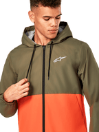 Alpinestars Speeder Windbreaker-Jacke, leichte Windjacke mit Kapuze, Colorblock-Design in Olivgrün und Military-Orange, weißes Alpinestars-Logo auf der Brust, schwarzer Reißverschluss und Kordelzüge, lässige, vom Motorsport inspirierte Sportbekleidung