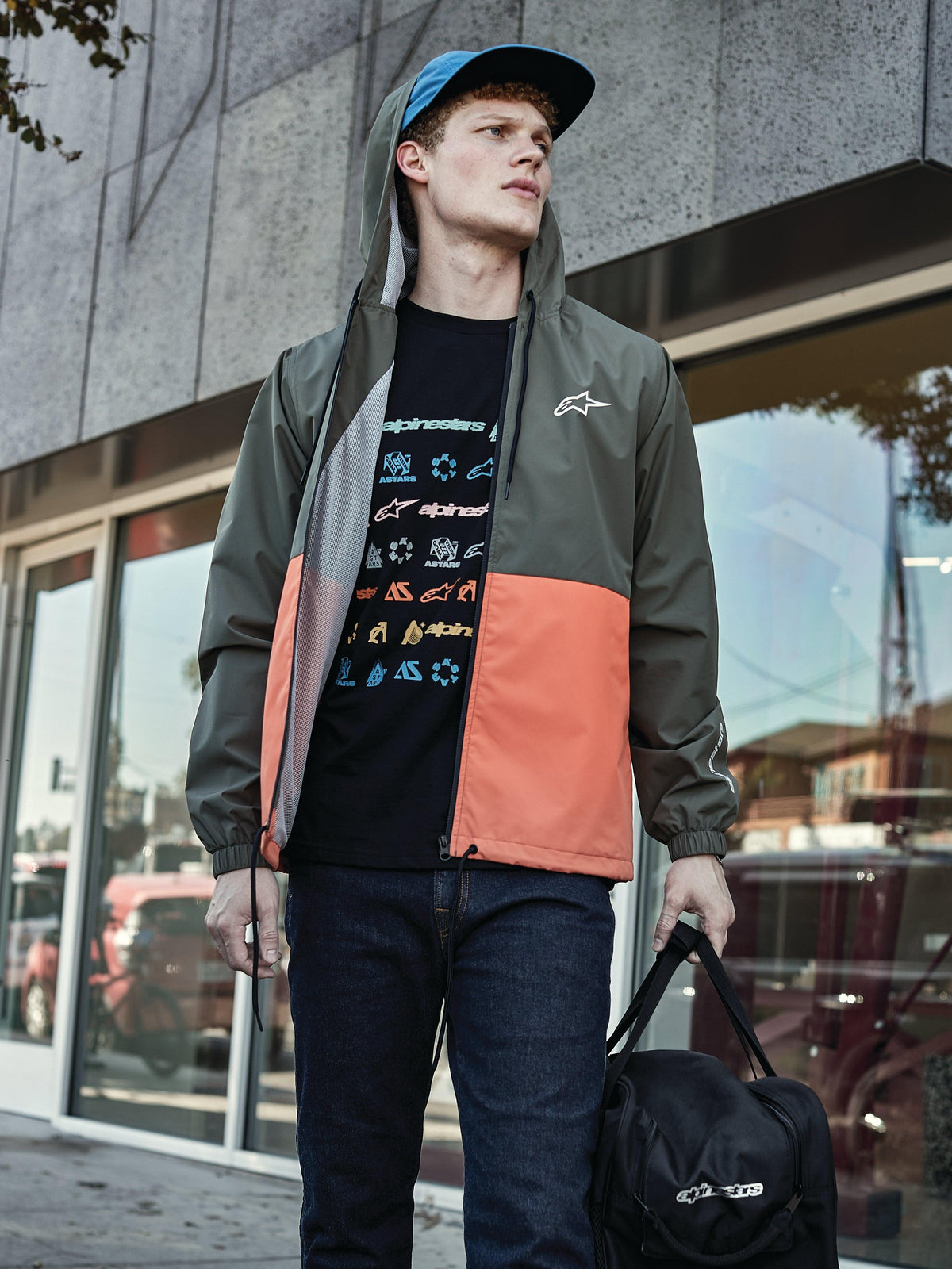 Mann in Alpinestars Speeder Windbreaker-Jacke in Military Orange, Color-Block-Design in Militärgrün und Orange, Kapuze mit Mesh-Futter, kombiniert mit schwarzem Grafik-Logo-T-Shirt, blauen Jeans und schwarzer Alpinestars-Sporttasche, lässiger Sportswear-Lifestyle-Look