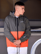 Mann in der Alpinestars Speeder Windbreaker-Jacke, Color-Block-Design in Military Orange und Dunkelgrün, lässige Kapuzenjacke mit weißen Logos auf Brust und Ärmel, kombiniert mit einer weißen Hose für einen vom Motorsport inspirierten Lifestyle-Look