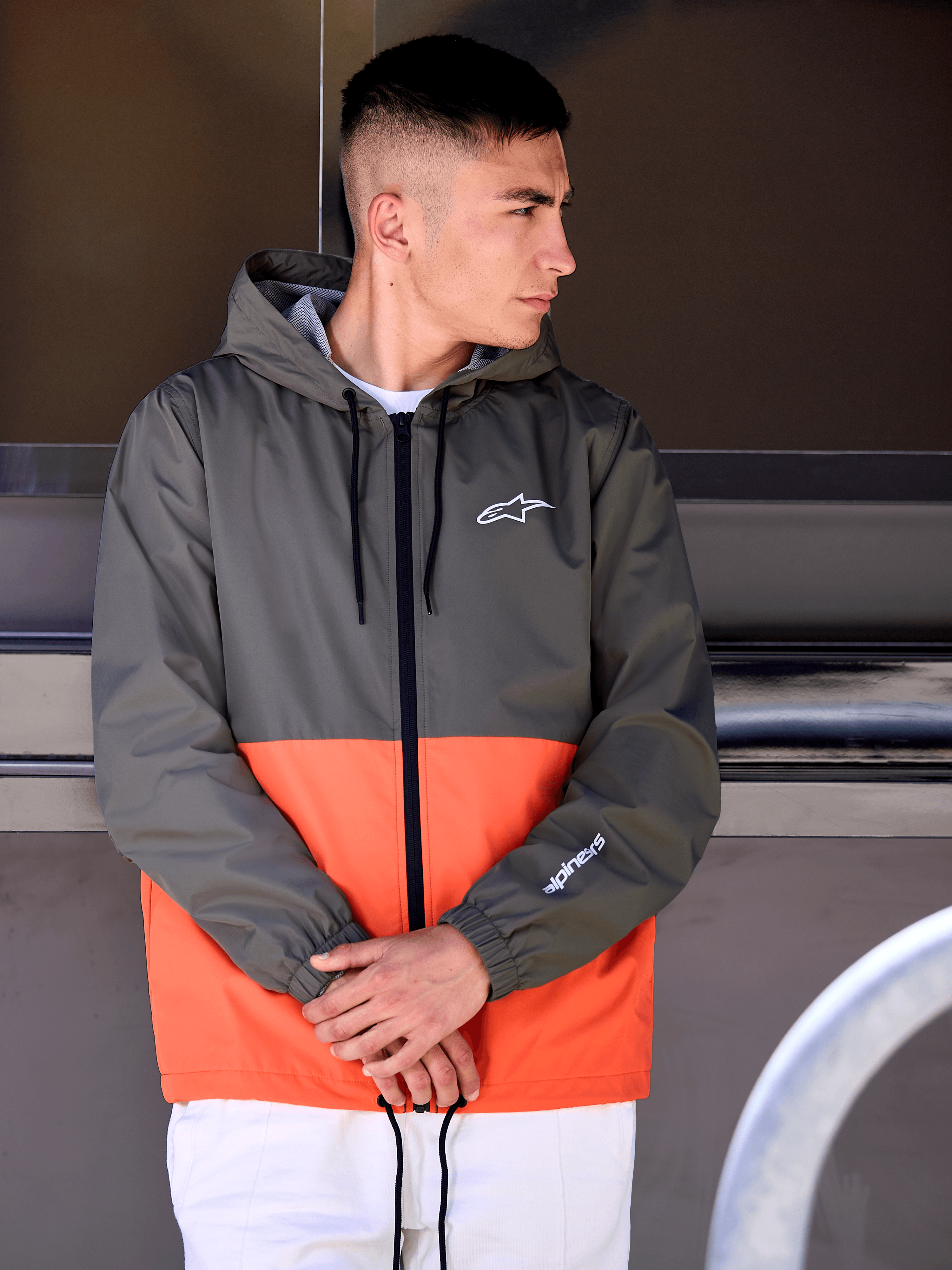 Mann in der Alpinestars Speeder Windbreaker-Jacke, Color-Block-Design in Military Orange und Dunkelgrün, lässige Kapuzenjacke mit weißen Logos auf Brust und Ärmel, kombiniert mit einer weißen Hose für einen vom Motorsport inspirierten Lifestyle-Look