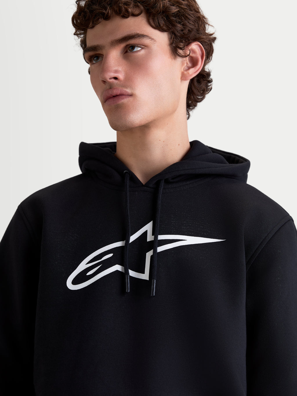 Alpinestars Ageless V3 Hoodie, Herren-Sportswear-Kapuzenpullover aus Fleece, Schwarz und Weiß, Detailansicht am Model mit großem weißem Astars-Logo auf der Brust und Kapuze mit Kordelzug.