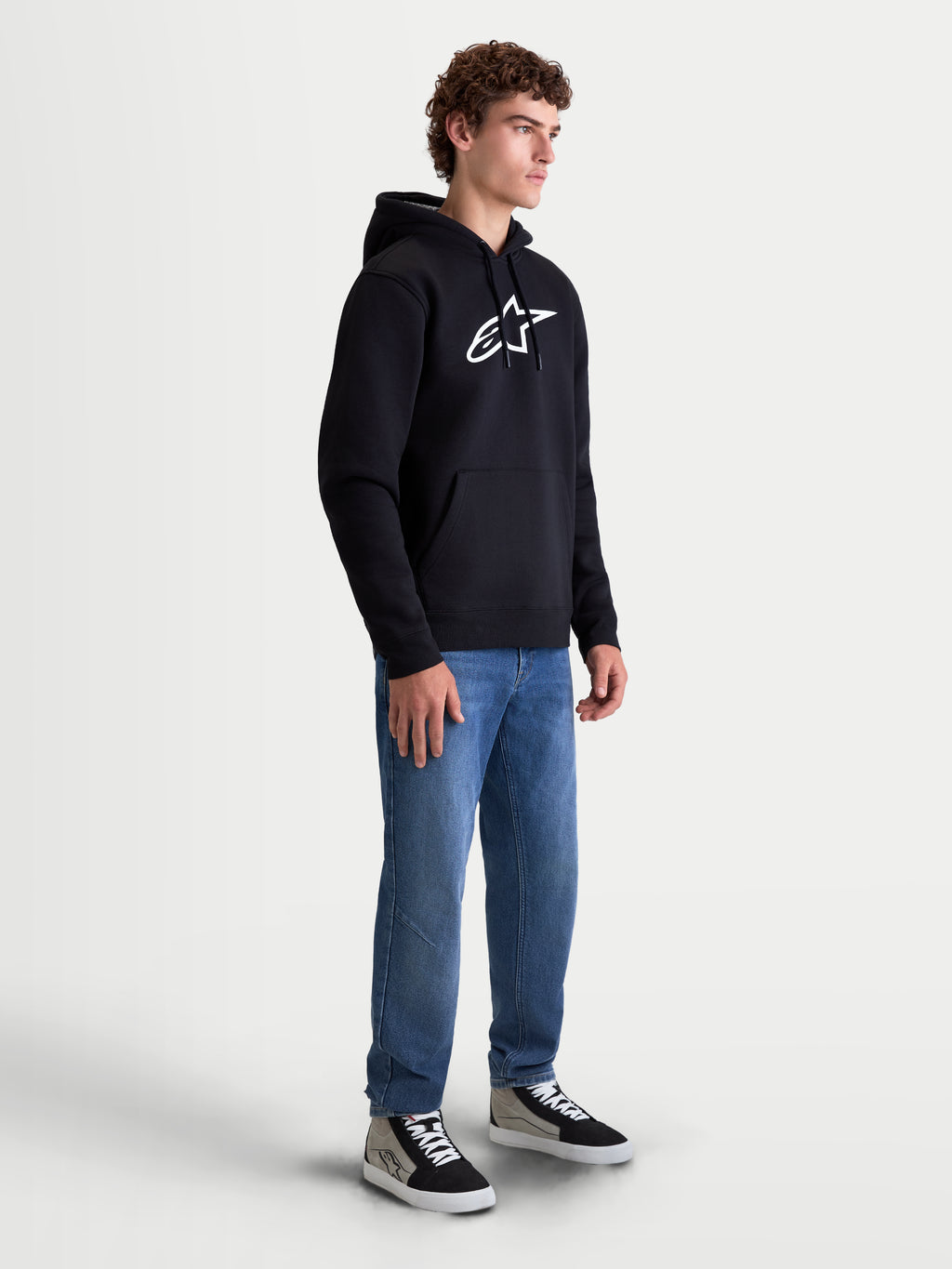 Alpinestars Ageless V3 Hoodie, Herren-Kapuzenpullover, Sportswear für Motorsport-Enthusiasten, Schwarz und Weiß, großes weißes gesticktes Logo auf der Brust, weiches Fleece-Material, Kapuze mit Kordelzug und Kängurutasche. Gezeigt an einem Model im Lifestyle-Kontext.