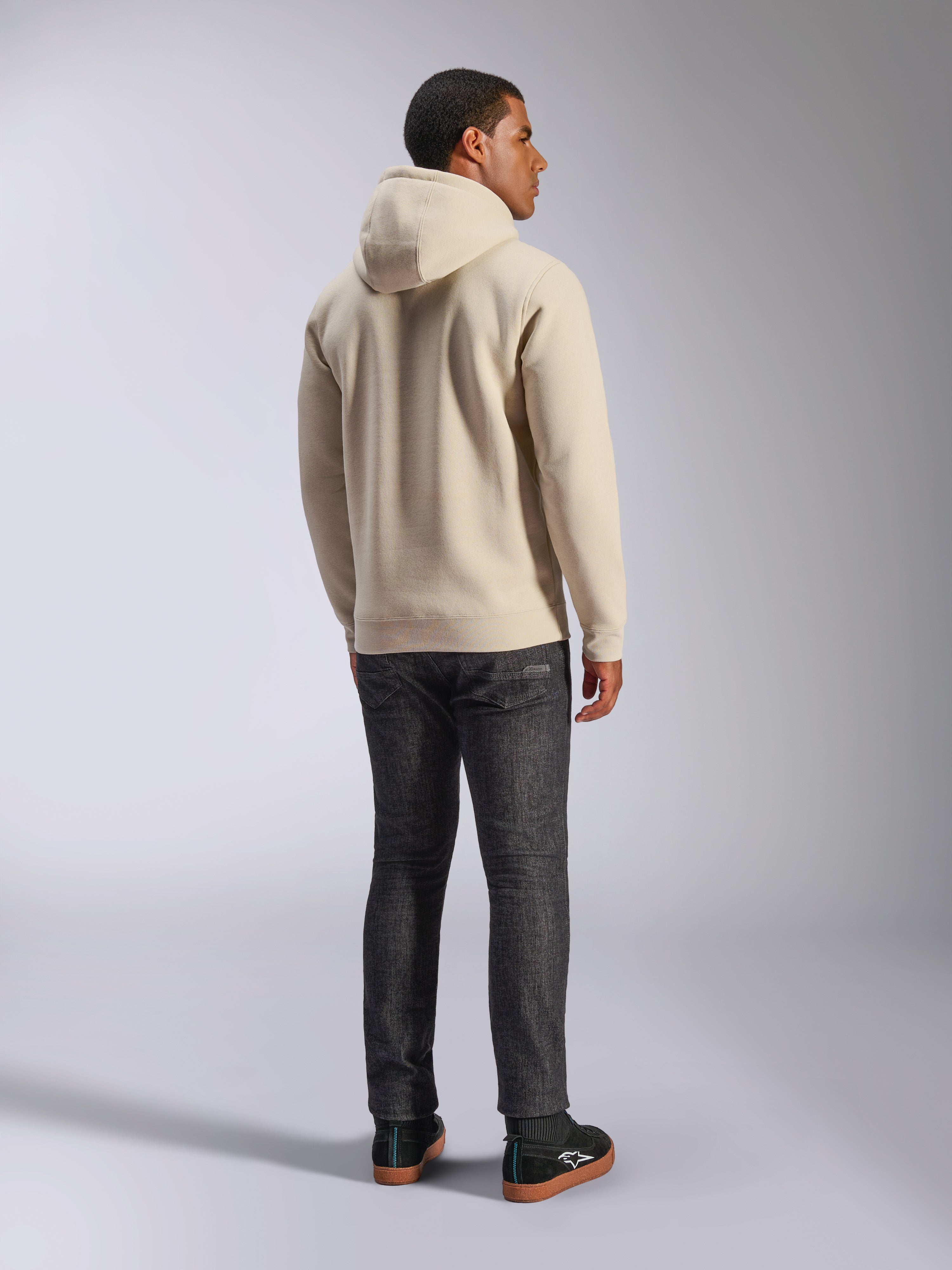 Alpinestars Ageless V3 Hoodie, lässiger Fleece-Kapuzenpullover in Off-White von hinten gezeigt, mit Rippbündchen an Ärmeln und Saum, getragen mit dunkler Slim-Fit-Jeans und schwarzen Alpinestars-Sneakern