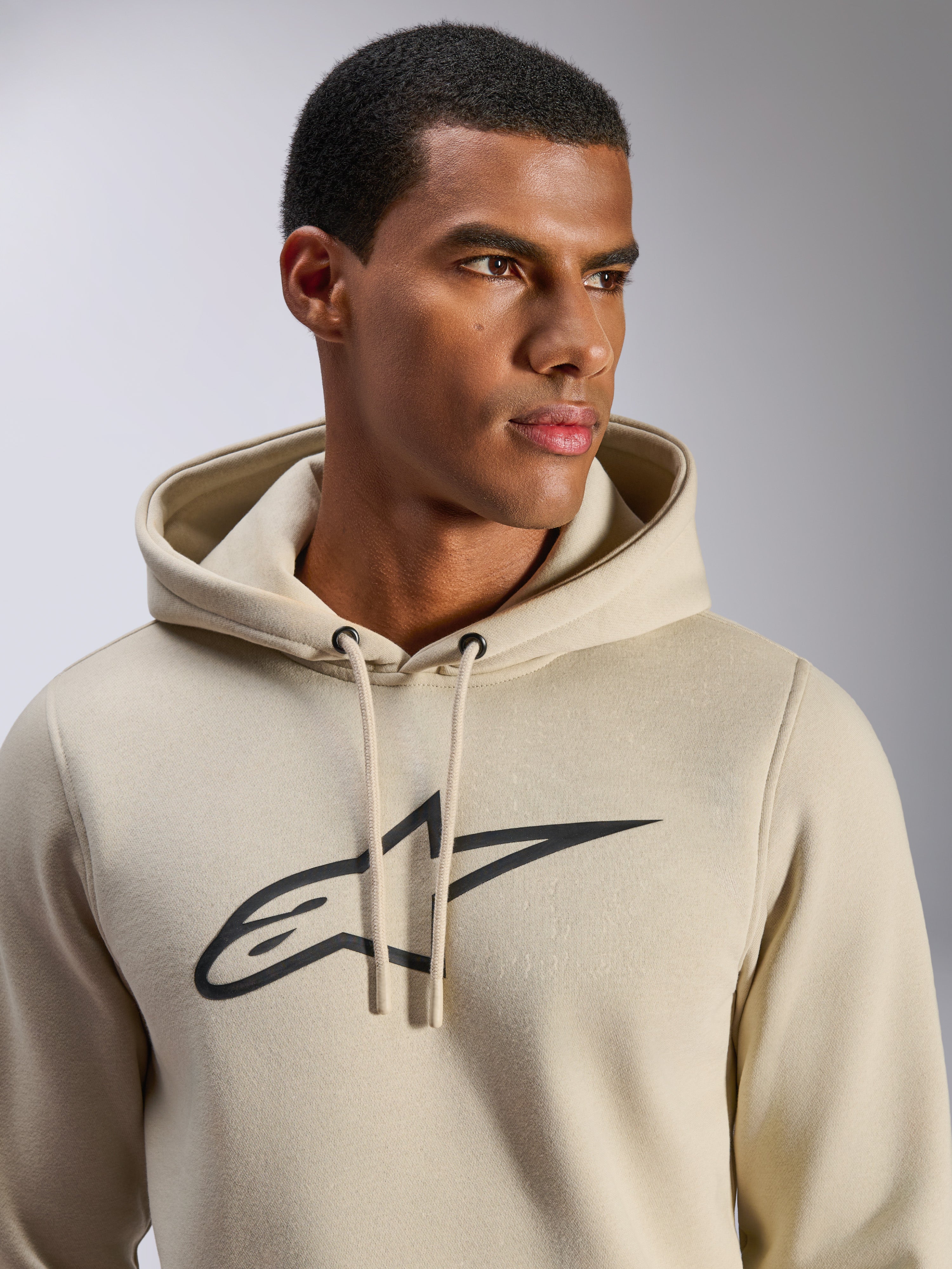 Alpinestars Ageless V3 Hoodie, lässige Sportbekleidung, Off-White mit schwarz besticktem Alpinestars Stern-Logo, Kapuze mit Kordelzug, Nahaufnahme am Model, weiches Fleece, modernes, vom Motorsport inspiriertes Design