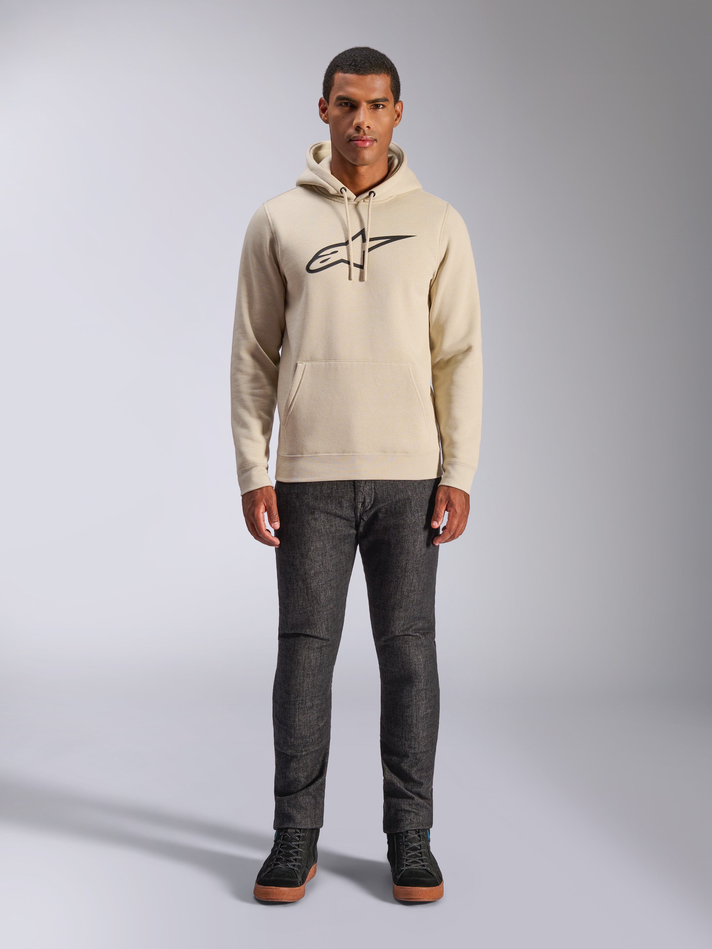 Alpinestars Ageless V3 Hoodie, lässige Sportbekleidung, Off-White mit schwarz gesticktem Logo, getragen von einem Mann mit dunkelgrauer Hose und schwarzen Sneakern, mit Kapuze mit Kordelzug und Kängurutasche, modernes vom Motorsport inspiriertes Design