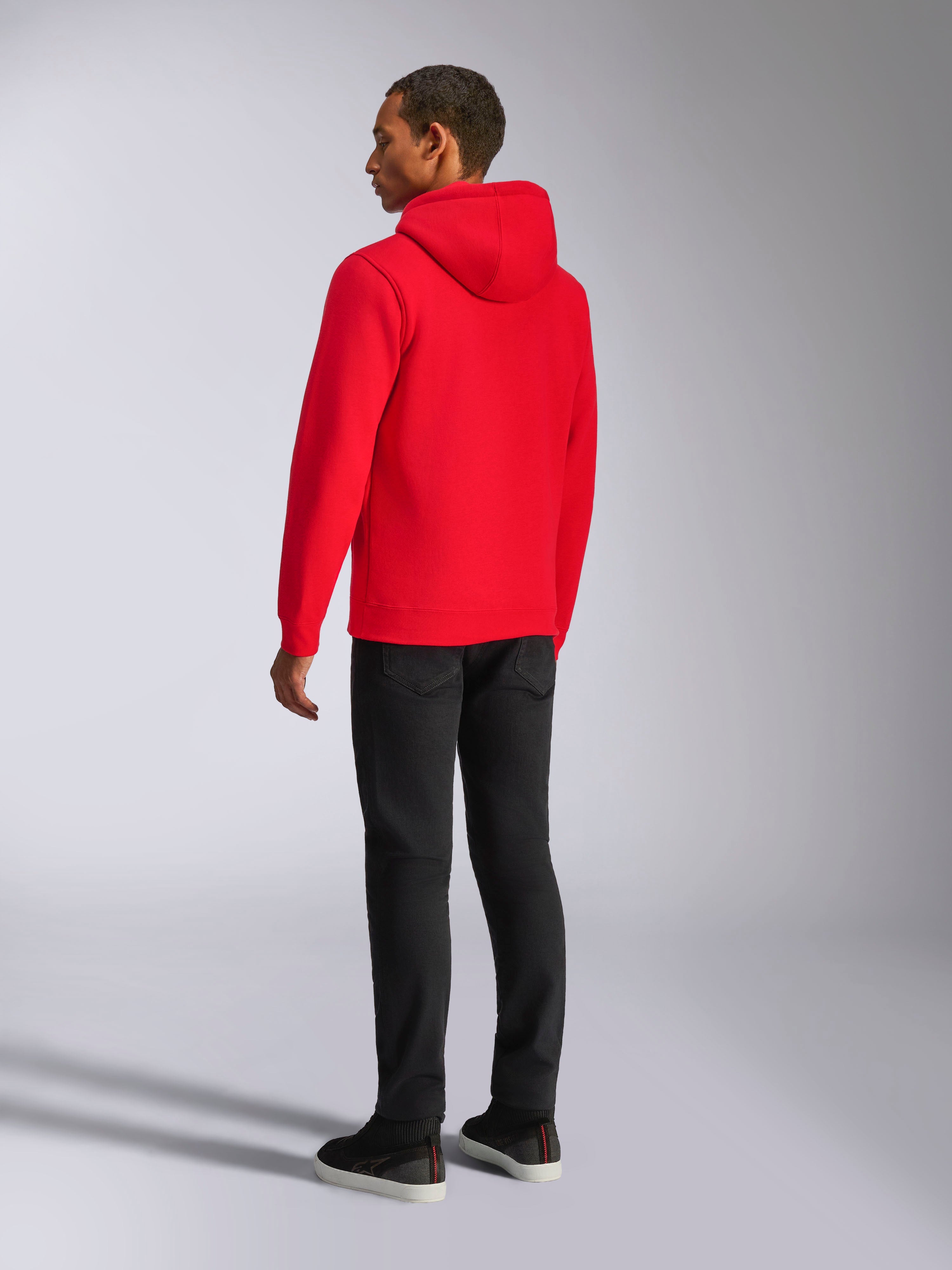 Alpinestars Ageless V3 Hoodie, lässige Streetwear, leuchtendes Rot, Rückansicht des Models, mit jerseygefütterter Kapuze, Rippbündchen an Ärmeln und Saum, kombiniert mit schwarzer Hose, modernes, vom Motorsport inspiriertes Design.