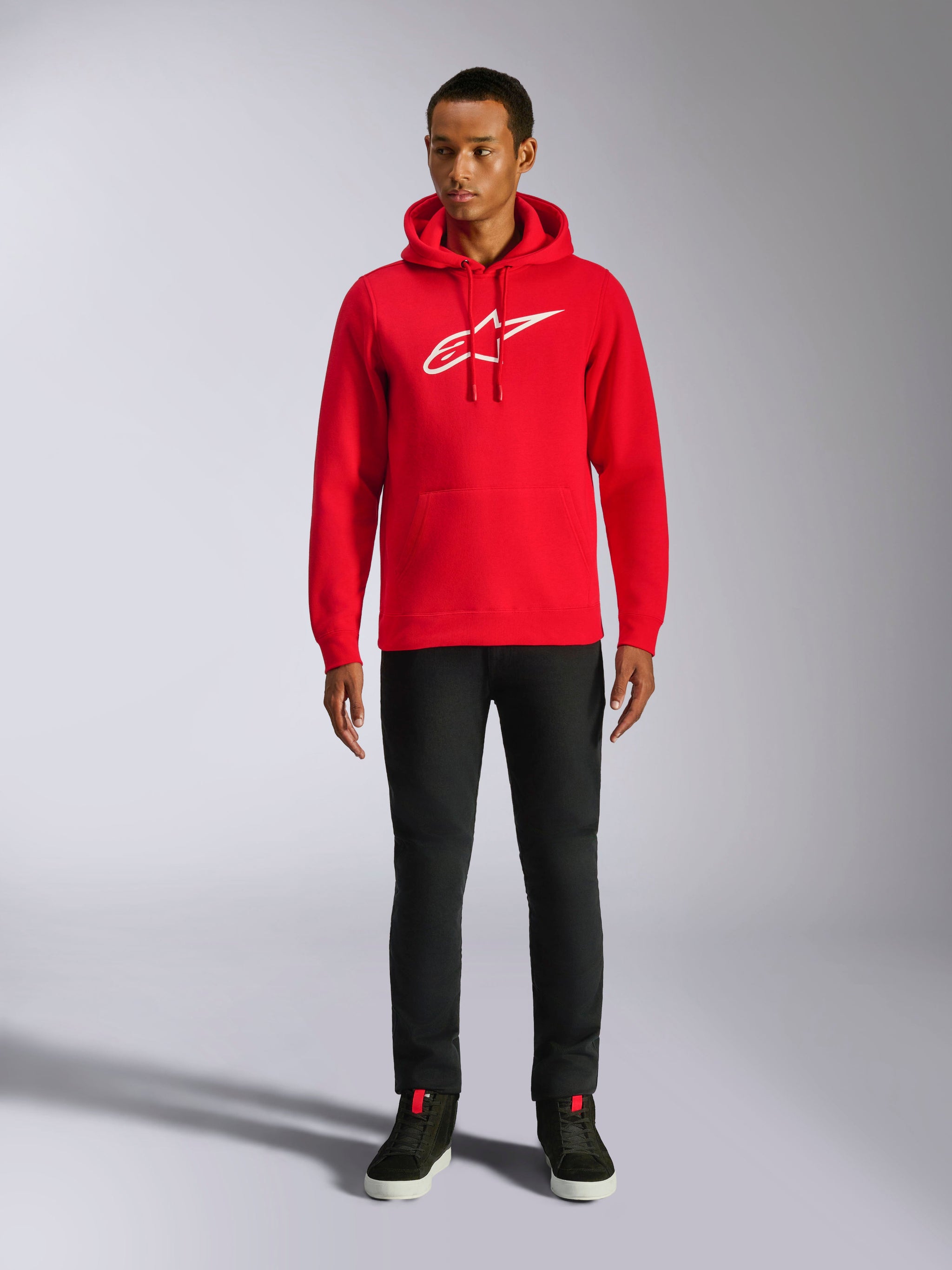 Alpinestars Ageless V3 Hoodie, lässige Streetwear, Rot mit weißem gesticktem Alpinestars-Logo, getragen von einem männlichen Model mit schwarzer Hose, Kapuze mit Kordelzug, Kängurutasche vorne, modernes, vom Motorsport inspiriertes Design