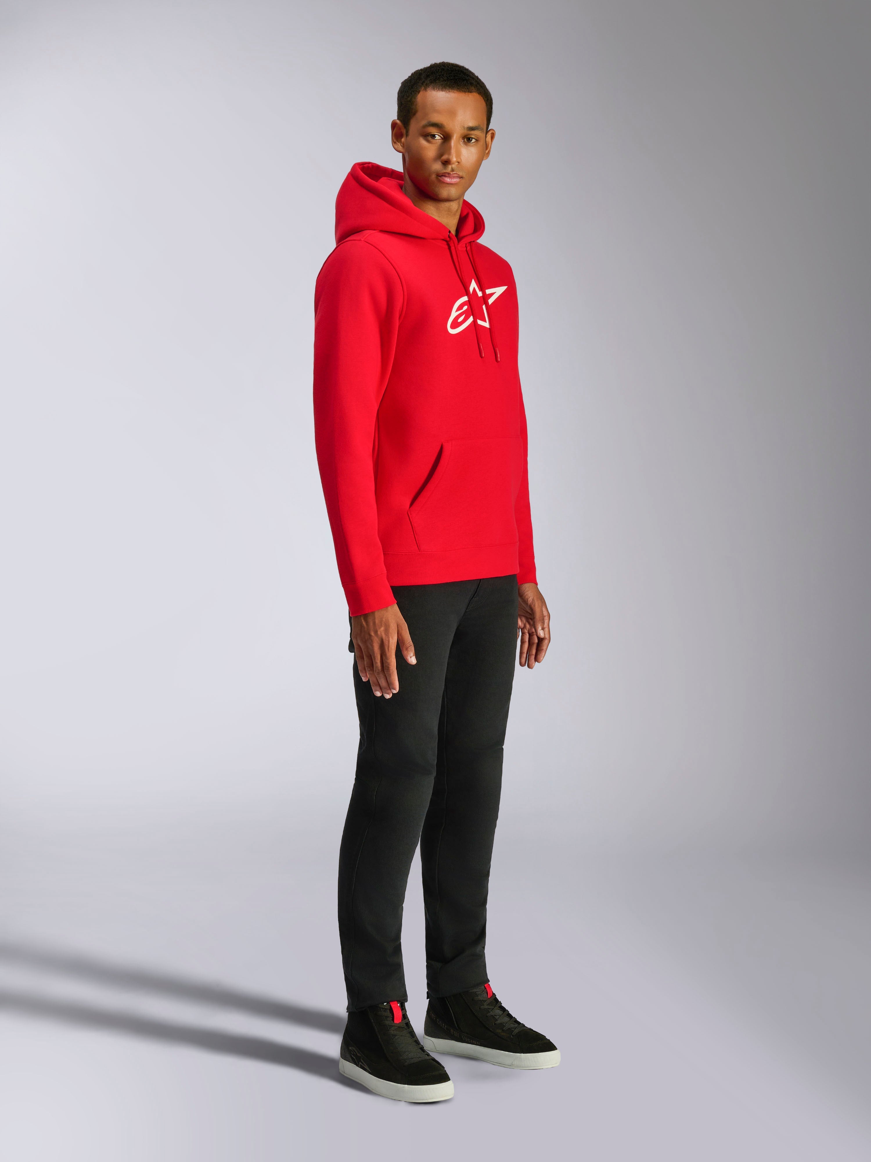 Alpinestars Ageless V3 Hoodie, Rot mit weiß gesticktem Logo, lässiger Sportswear-Hoodie, getragen von einem Mann mit schwarzer Hose und schwarzen Sneakern, mit Kapuze mit Kordelzug und Rippbündchen