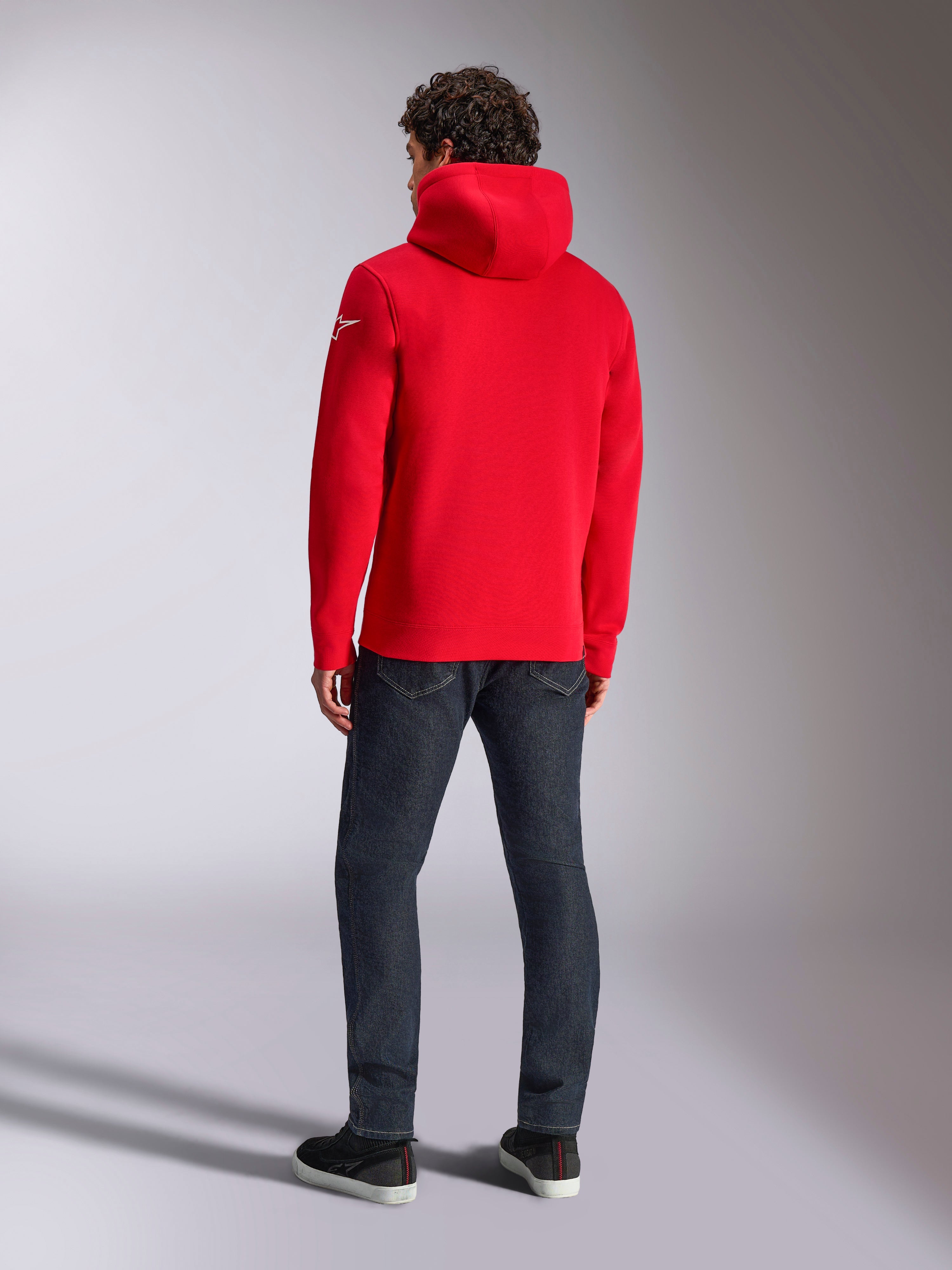 Alpinestars Linear Wordmark V3 Hoodie, lässiger roter Kapuzenpullover, Rückansicht mit Kapuze und weißem Ageless-Logo am Ärmel, kombiniert mit dunkelblauer Jeans und schwarzen Schuhen, Lifestyle-Motorsport-Streetwear