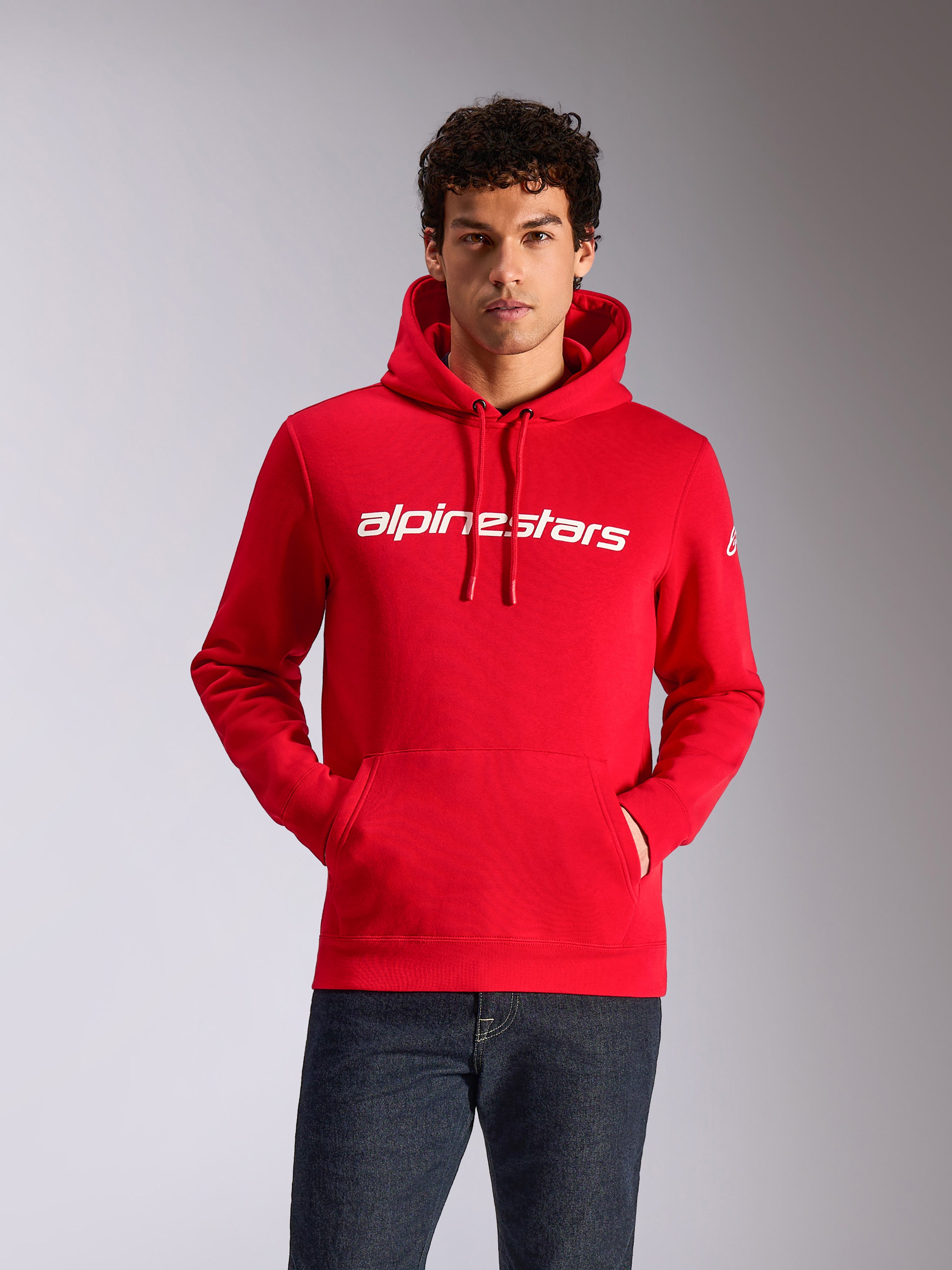 Alpinestars Linear Wordmark V3 Hoodie, Rot mit weißem Wortmarken-Logo, lässige Streetwear, getragen von einem Model mit Händen in der Kängurutasche, Kapuze mit Kordelzug, dunkle Denim-Jeans, modernes, vom Motorsport inspiriertes Design