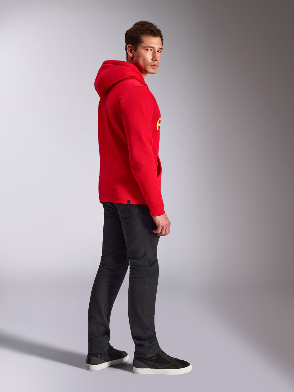 Alpinestars Linear Wordmark V3 Hoodie, Rot, lässige Streetwear, Modell in der Seiten- und Rückansicht trägt einen roten Kapuzenpullover mit gelbem Branding, dunkelgraue Jeans und schwarze Sneaker, Studio-Lifestyle-Aufnahme