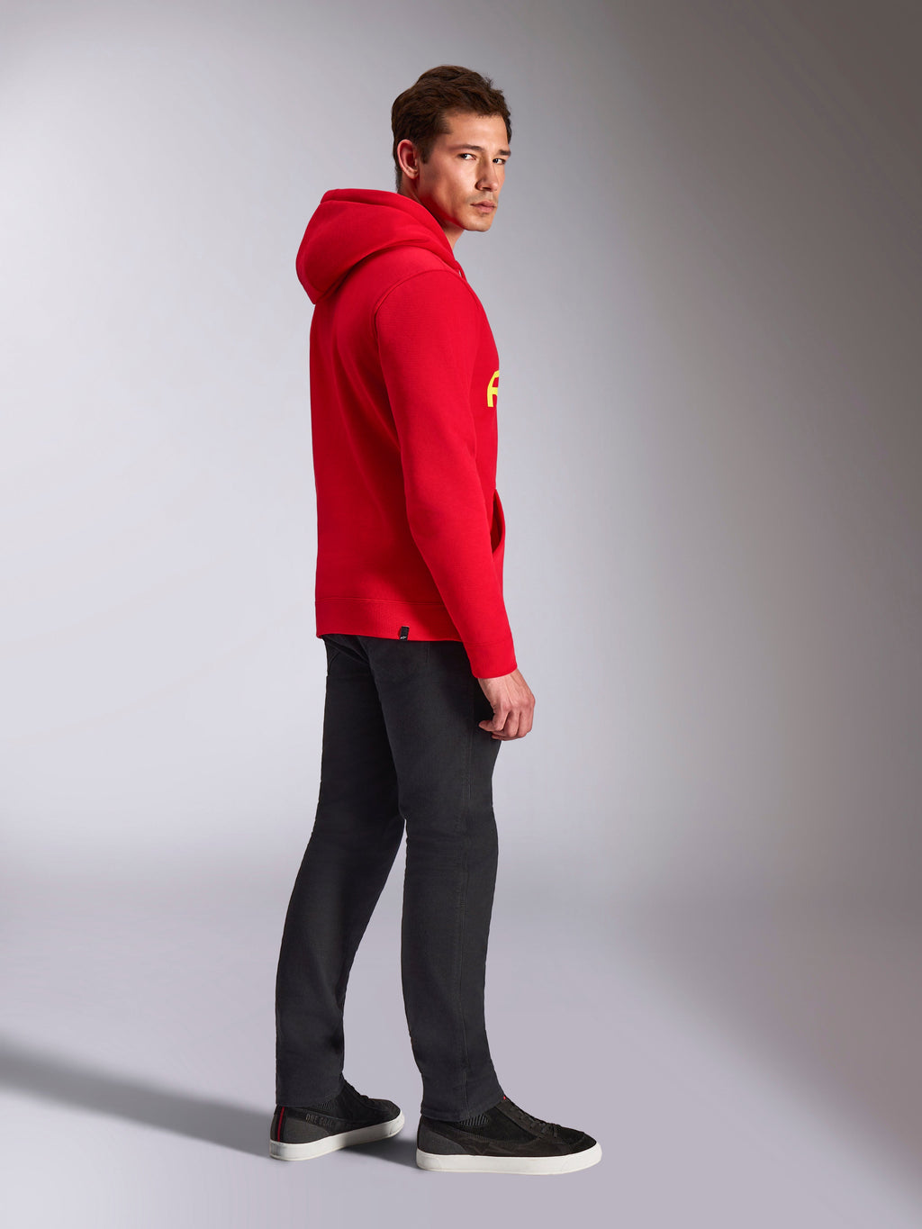 Alpinestars Ride V3 Hoodie, lässige Sportbekleidung, Rot mit Hi-Vis Gelbem Branding, männliches Model trägt roten Hoodie mit schwarzer Hose und Sneakern, Seitenprofil, Fleece-Pullover-Design mit Rippbündchen an Ärmeln und Saum.
