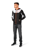 Alpinestars Sessions V3 Hoodie, lässige Kapuzenjacke mit Reißverschluss, Schwarz, Weiß und Rot, getragen von einem Model mit dunkler Jeans und schwarzen Sneakern, mit weißen Color-Block-Einsätzen an Brust und Ärmeln, rotem Reißverschluss und Kordelzug, großem Brustlogo, Poly-Fleece, vom Motorsport inspirierte Streetwear