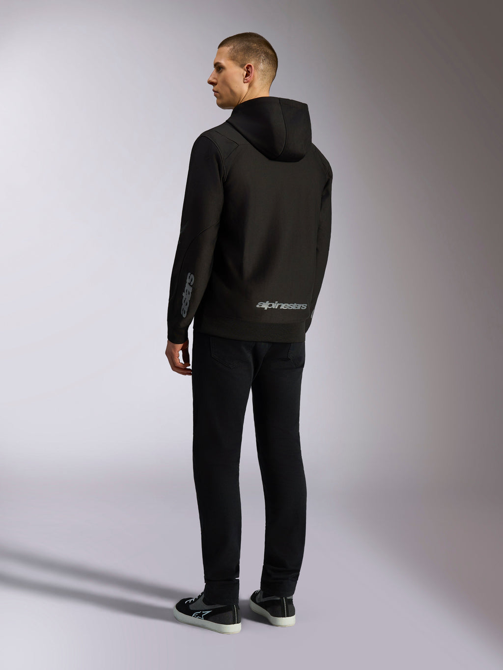 Alpinestars Sessions V3 Hoodie, schwarze Casual-Streetwear, Rückansicht an einem Model, mit grauem Alpinestars-Schriftzug am unteren Rücken und Astars-Logo auf dem Ärmel, Kapuzenjacke mit Reißverschluss und modernem, vom Motorsport inspiriertem Design