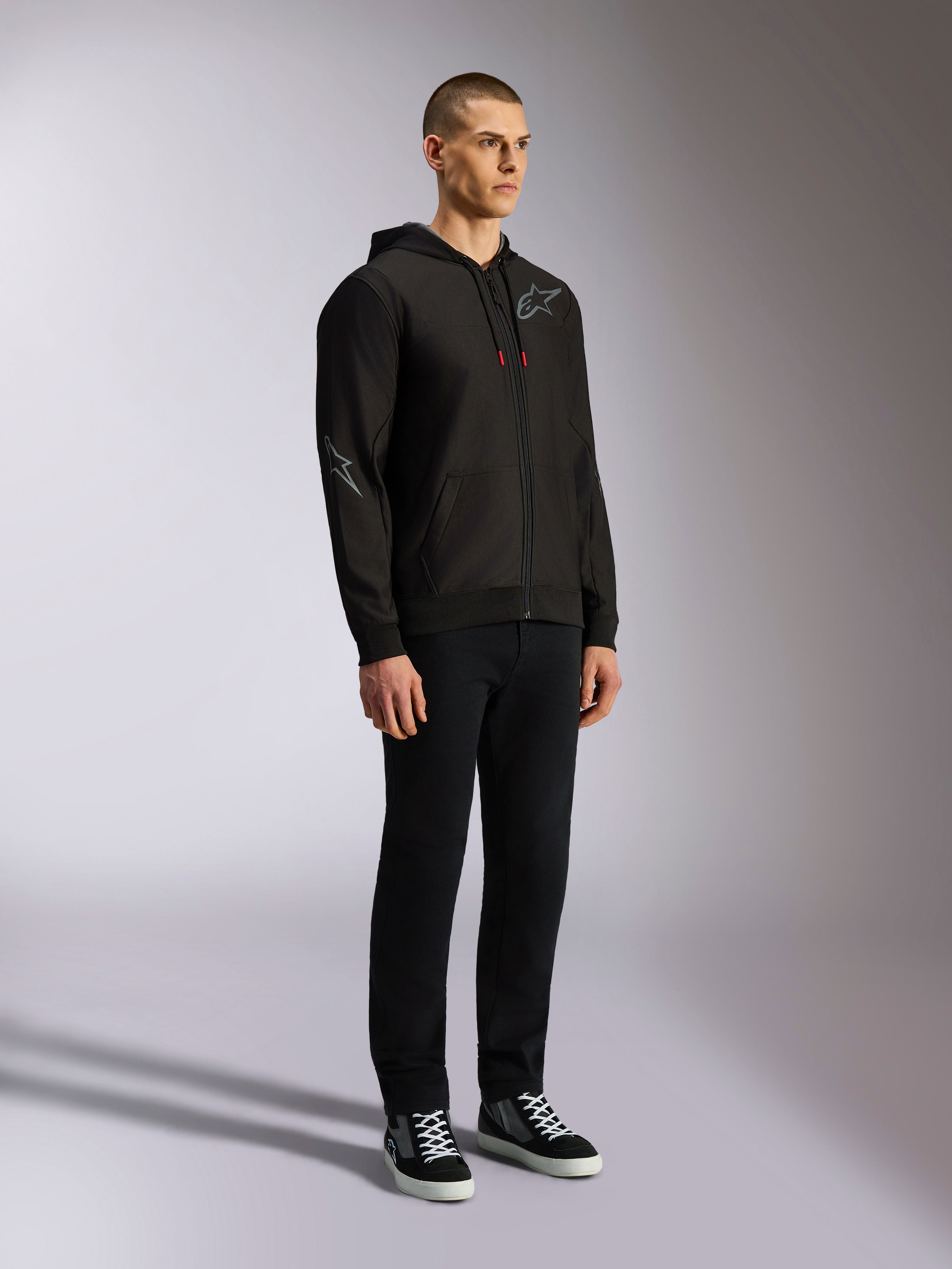 Alpinestars Sessions V3 Hoodie, lässige Motorsport-Streetwear, schwarze Kapuzenjacke mit Reißverschluss, grauem Stern-Logo auf Brust und Ärmel, Kordelzügen mit roten Enden, Rippbündchen und Saum, getragen von einem Model mit schwarzer Hose und Sneakern.