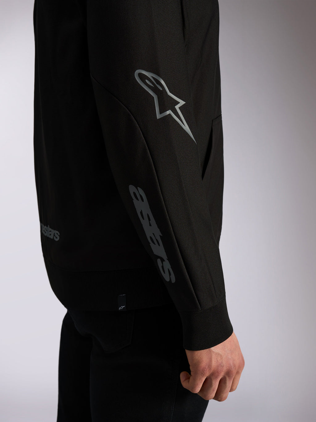Alpinestars Sessions V3 Hoodie, schwarze Casual-Streetwear, Detailaufnahme des Ärmels mit grauem Astars-Logo am Ellbogen und Wortmarke am Unterarm, Rippbündchen, vom Motorsport inspiriertes Design