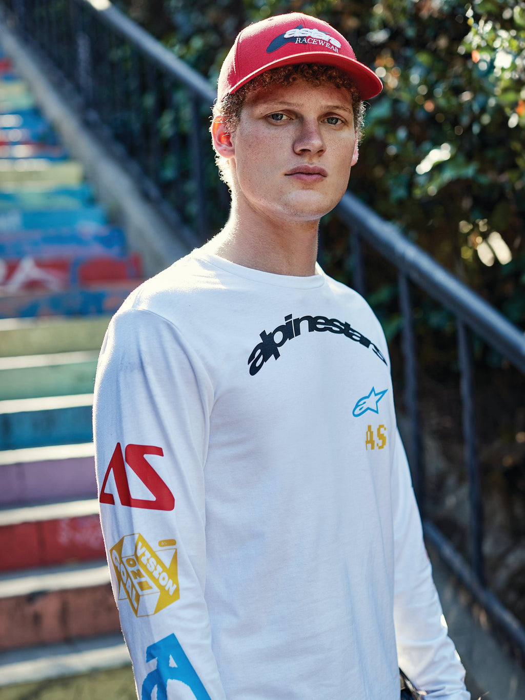 Alpinestars Brundo CSF Langarmshirt, Weiß, mit schwarzem Branding auf der Brust und mehrfarbigen Grafiken an den Ärmeln, getragen von einem Mann mit roter Cap, lässiger Motorsport-Streetwear-Stil.