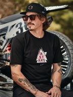 Alpinestars Gambit Trucker Mütze in Schwarz mit kreisförmigem Logo-Patch, getragen mit Alpinestars Soloist SS T-Shirt in Schwarz, Lifestyle-Aufnahme eines Mannes mit Sonnenbrille und Tätowierungen vor einem Motocross-Motorrad.