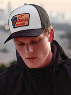 Alpinestars Hardware Trucker Hat in Military White, getragen von einem Mann in einem schwarzen Hoodie, weißes Frontpanel mit schwarzem gebogenem Schirm und schwarzer Mesh-Rückseite, beiger Stickereipatch mit rotem Alpinestars Motocross-Branding, lässige Lifestyle-Motorsport-Streetwear