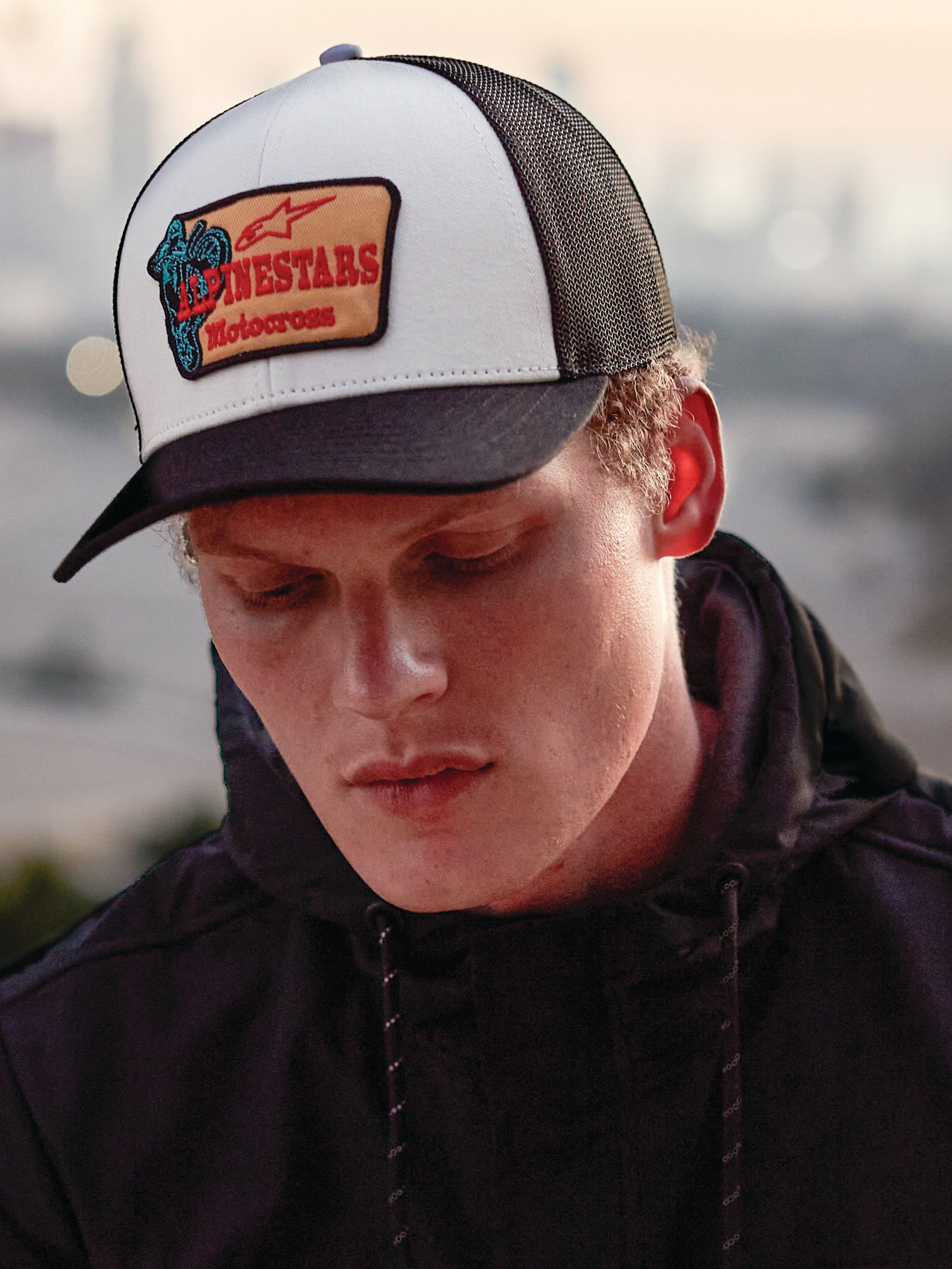 Alpinestars Hardware Trucker Hat in Military White, getragen von einem Mann in einem schwarzen Hoodie, weißes Frontpanel mit schwarzem gebogenem Schirm und schwarzer Mesh-Rückseite, beiger Stickereipatch mit rotem Alpinestars Motocross-Branding, lässige Lifestyle-Motorsport-Streetwear