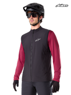 Alpinestars A-Dura Thermoweste, schwarze Mountainbike-Thermoweste für Herren, getragen von einem Model mit Helm und rotem Langarm-Trikot, isoliert für MTB-Fahrten bei kaltem Wetter